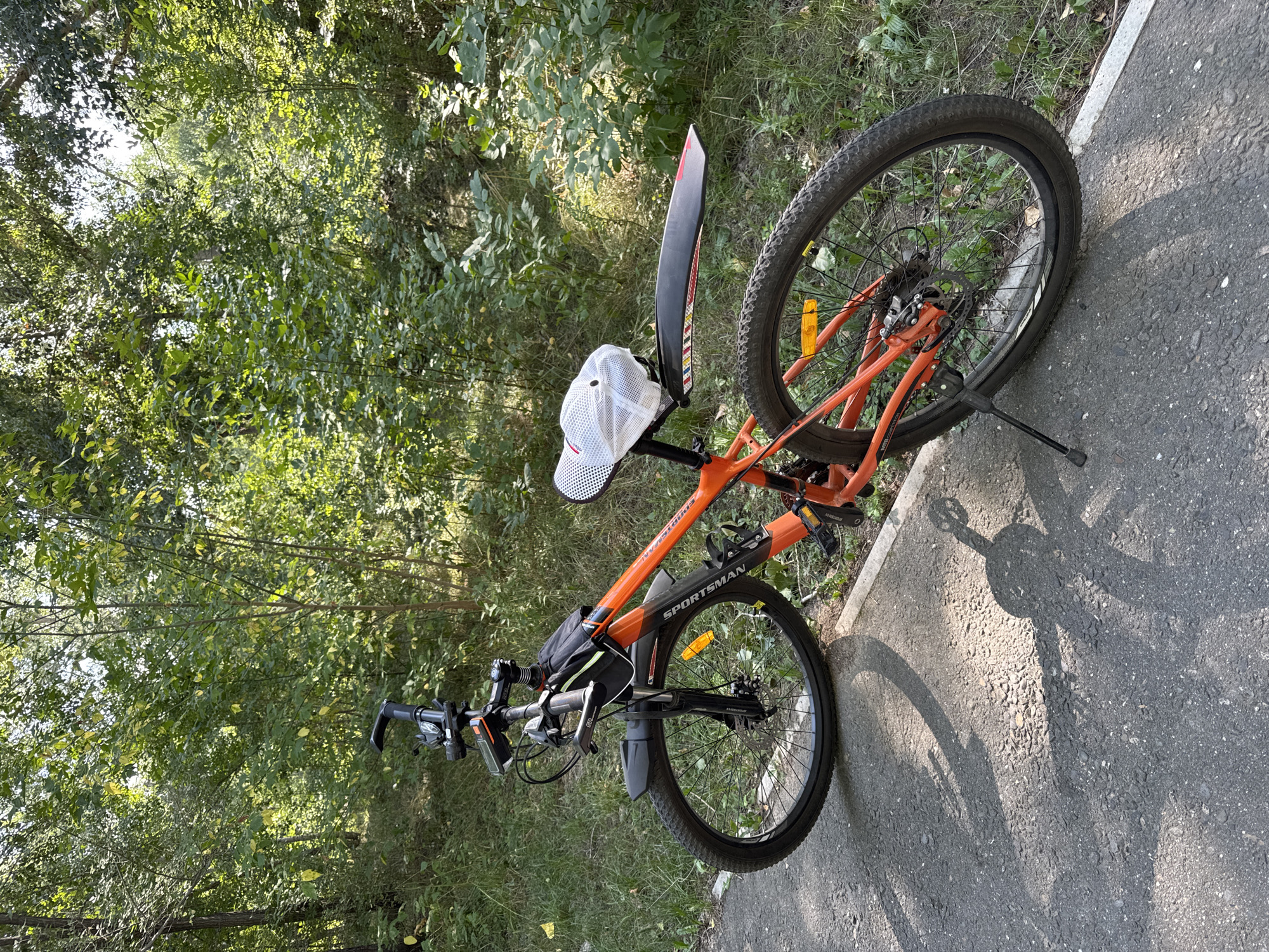 An orange mountain bike is parked on a paved path next to a grassy area with trees. The bike is leaning on its kickstand. It has a black fender on the rear wheel and a black bag attached to the handlebars. A white cap with a checkered pattern is placed on the seat. The background consists of lush green foliage and trees, suggesting an outdoor, possibly park-like setting. The scene is illuminated by bright sunlight, casting shadows on the path. The specific location in Oskemen, Kazakhstan, is not identifiable from the visual information present in the image. There are no people or animals visible in the image.