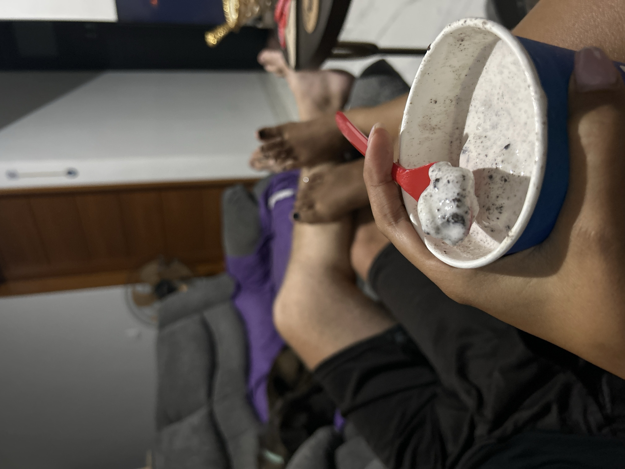 In a dimly lit indoor setting, a close-up shot focuses on a hand holding a cup of Cookies and Cream ice cream with a red spoon. The ice cream appears to be a vanilla base with dark cookie crumbles throughout. In the background, several people's legs and feet are visible, suggesting a gathering or shared activity. One person is wearing black shorts, while another has on purple and grey clothing. The feet in the background show painted toenails and some jewelry, indicating personal adornment. The overall mood seems relaxed and casual, possibly a moment of shared enjoyment of dessert. The time of day is unclear, but the ambient lighting suggests it could be evening or indoors during the day. The presence of furniture like a couch indicates a domestic or casual social environment. No visible text is present in the image.