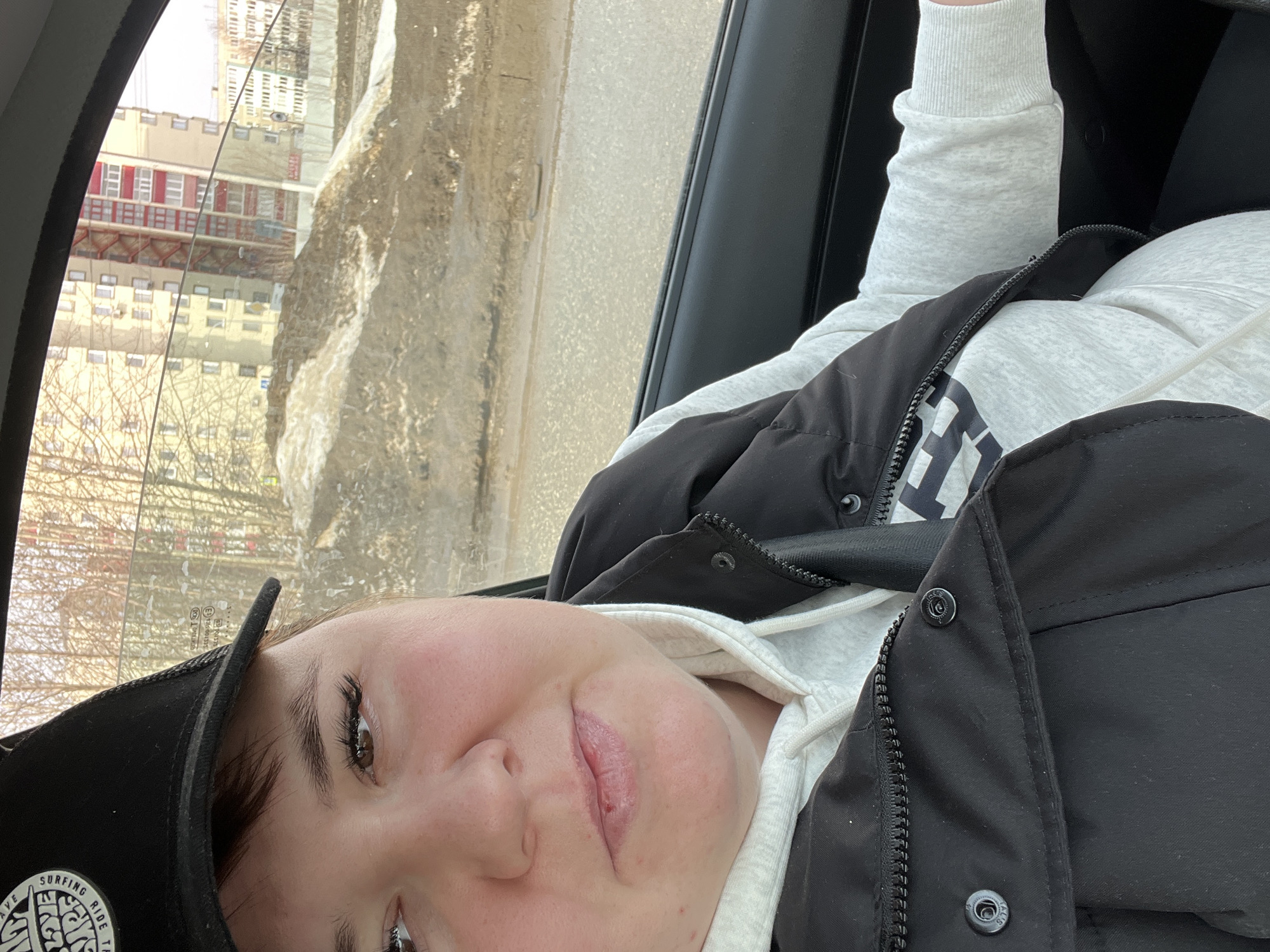 A female individual is depicted in the driver's seat of a vehicle, captured in a close-up selfie-style photograph. She wears a black baseball cap with white text on the front, partially obscuring her dark brown hair. Her eyes are brown with prominent dark eyelashes, and her lips are slightly parted, revealing a small red mark or scab on the lower lip. She is dressed in a light grey hooded sweatshirt, with a black sleeveless puffer vest or jacket worn over it. A seatbelt strap is visible across her chest.
Through the vehicle's passenger-side window, an urban environment in Chaikovsky, Russia, is visible, suggesting a late winter or early spring setting. The immediate foreground outside the window shows a large mound of dirt and debris partially covered with melting snow and ice, indicative of ground disturbance or construction. Beyond this, a wet, dark road surface is visible. In the background, multiple multi-story residential buildings are present. A prominent light yellow/beige apartment building features red-colored balconies or external stairwells. Bare deciduous trees are scattered throughout the scene. The overall lighting suggests daylight. Mirrored text, possibly "TOC 503" and "SANSISOS-3EA," is reflected on the vehicle's side window.