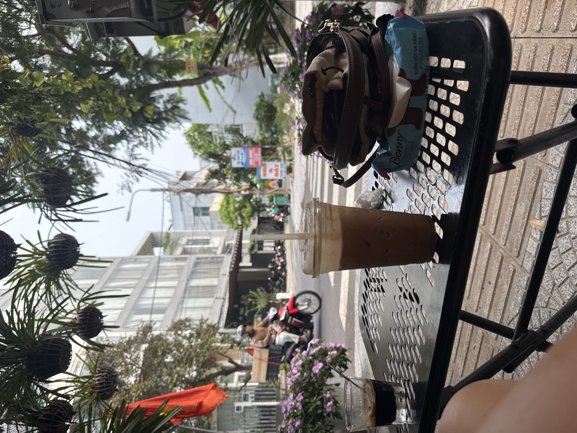 A medium-close shot angled toward a black metal table at an outdoor cafe in Đà Nẵng, Vietnam. On the table are a clear plastic cup of iced coffee with a straw, a brown purse, and a packet of wet wipes. In the blurry background, buildings line a street where scooters and a motorcycle drive by. Vegetation hangs from the building above. A short glass of coffee and an orange awning are on the table, close to the lower edge of the frame. Part of a person's leg is visible in the lower right corner.
