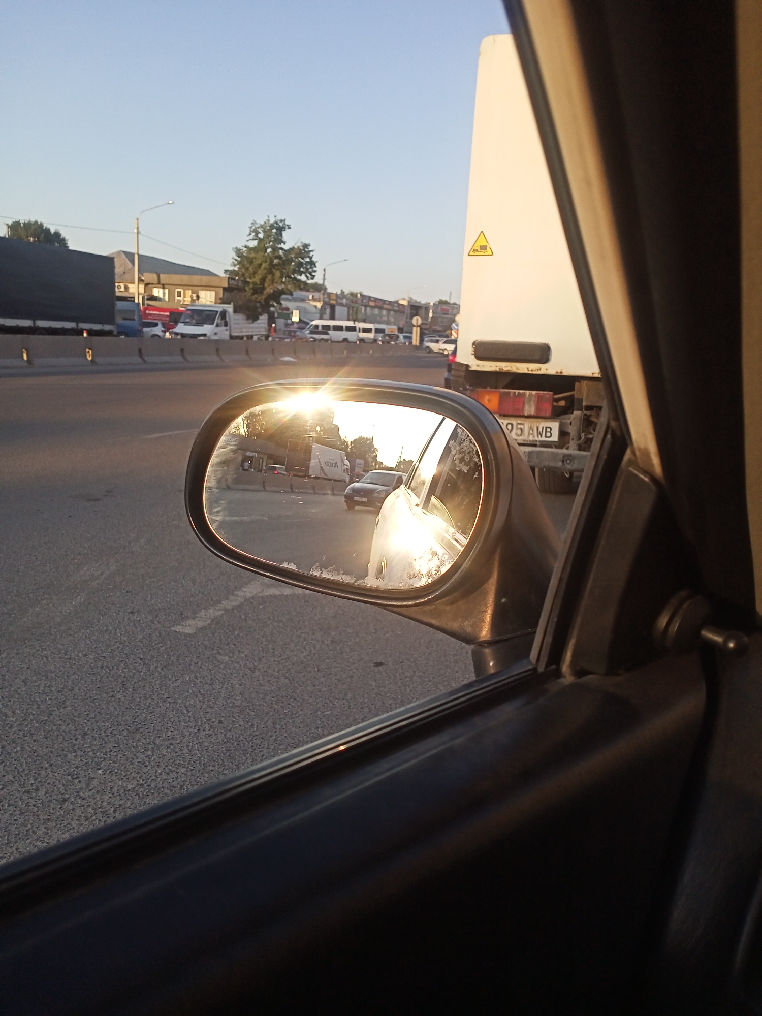 The image shows a view from inside a vehicle looking out at a street scene in Sokuluk, Kyrgyzstan. The sun is setting, casting a warm glow and creating reflections in the side-view mirror. In the mirror, we can see traffic, including a truck with a license plate that reads "05 AWB". The truck is white with a yellow triangular hazard sign. Beyond the mirror, the road is visible, with a concrete barrier and other vehicles. The sky is clear and blue, with some trees and buildings in the background. The overall impression is of a busy street during golden hour.