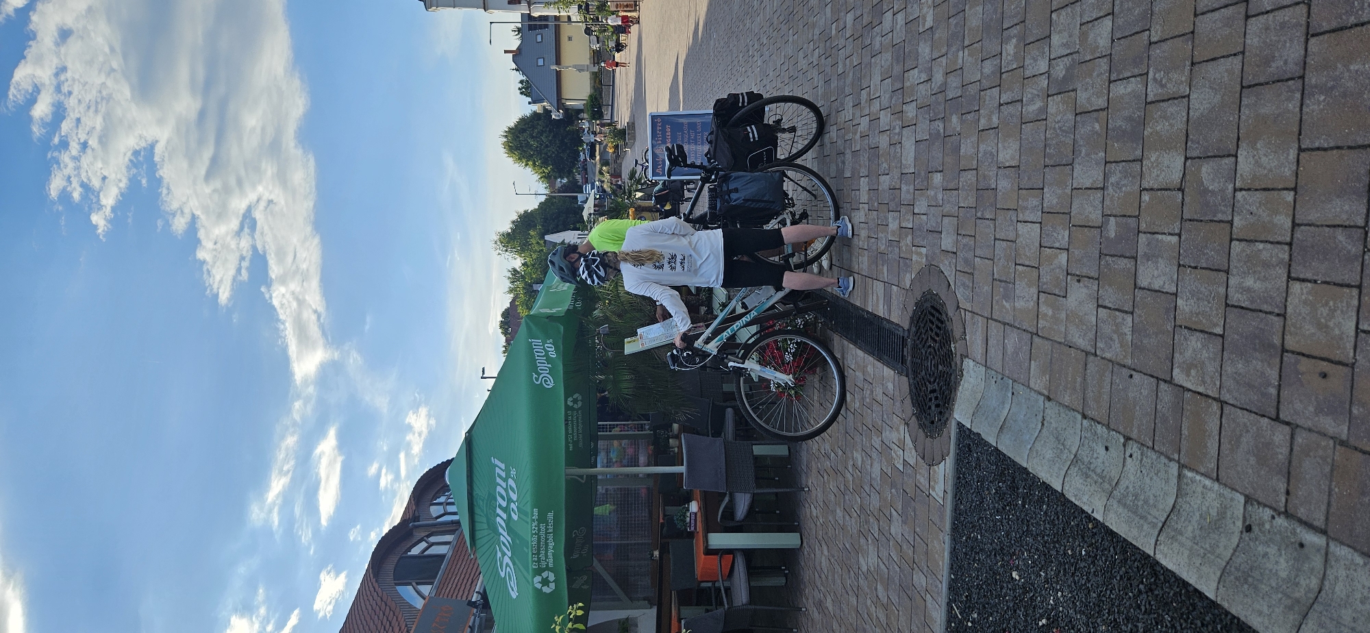 Two people, a woman and a man, are standing next to their bicycles on a paved area with a patterned brick surface. The woman, wearing a white long-sleeved shirt and black shorts, is holding a map or paper. The man, in a bright green shirt and helmet, is also looking at the paper. Both are wearing helmets, suggesting they are cyclists. Their bicycles are equipped with panniers, indicating they might be on a tour or journey.
Behind them, a green umbrella with the "Soproni 0.0%" logo is providing shade over an outdoor seating area with tables and chairs. The umbrella also displays text about the product being made from recycled plastic. A sign board with the word "ANGEBOT" (German for "offer") is visible behind the bicycles.
The scene is set in an outdoor environment with buildings and trees visible in the background. The sky is blue with scattered white clouds, suggesting it is daytime, likely late afternoon given the soft light. The overall impression is a break during a cycling trip, possibly at a cafe or rest stop. The location context indicates this is in Balatongyörök, Hungary.
