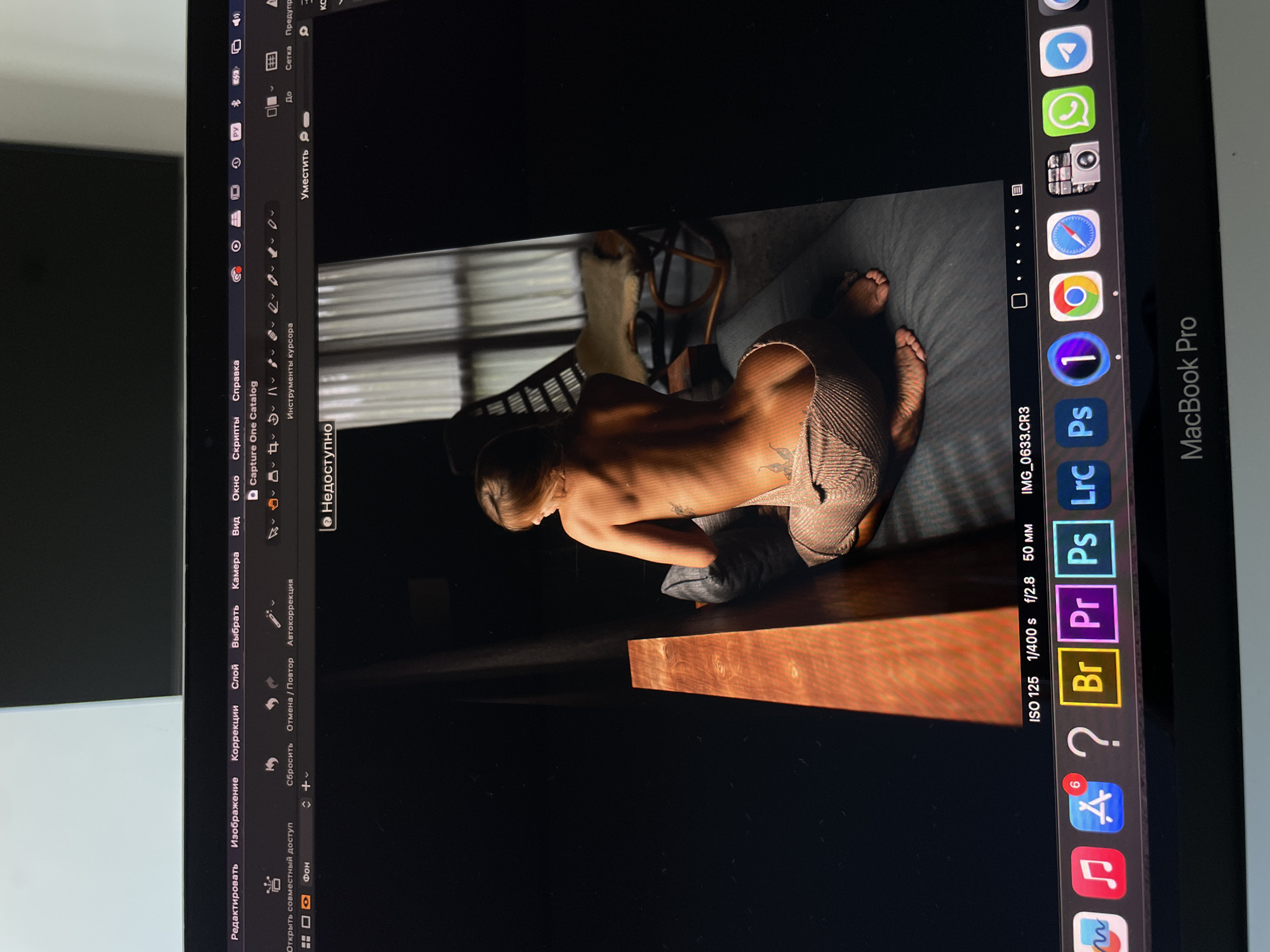 The image is a photograph of a MacBook Pro screen, displaying a partially visible photograph within the Capture One photo editing software. The edited photo depicts a woman kneeling on a bed or soft surface. The photo is taken from a low angle, possibly with a light source from the top left. The photo on the screen appears to be a low-light setting with the woman's back exposed and a small tattoo visible.
The MacBook Pro's dock is visible on the right side of the screen, and various toolbars and menus of Capture One are along the top and left edges. The parameters of the photo displayed in Capture One are ISO 125, 1/400s shutter speed, f/2.8 aperture, and a 50mm focal length.
There is no indication of a specific location such as Kazan, Russia, in the information available in the image.