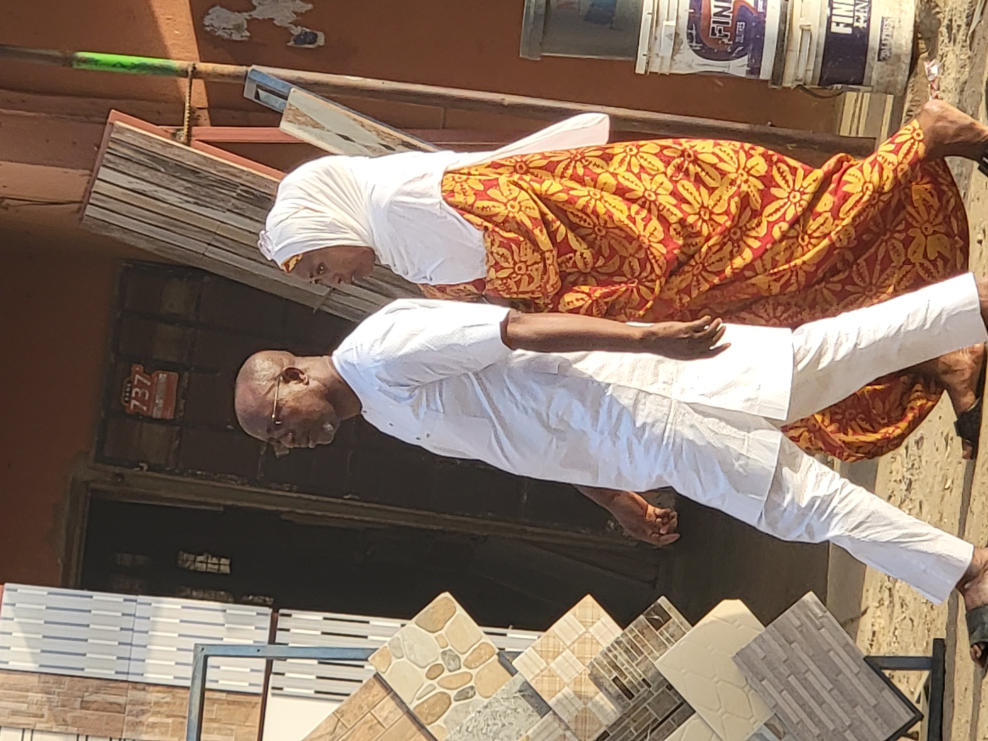 In Maiburiji, Nigeria, a woman in a white headscarf and a red and yellow floral print dress walks alongside a man dressed in a white shirt and trousers. They are walking on a sidewalk next to a building with a display of tiles. Behind them, there are paint buckets stacked on top of each other. A sign with the number "737" is visible on the building.