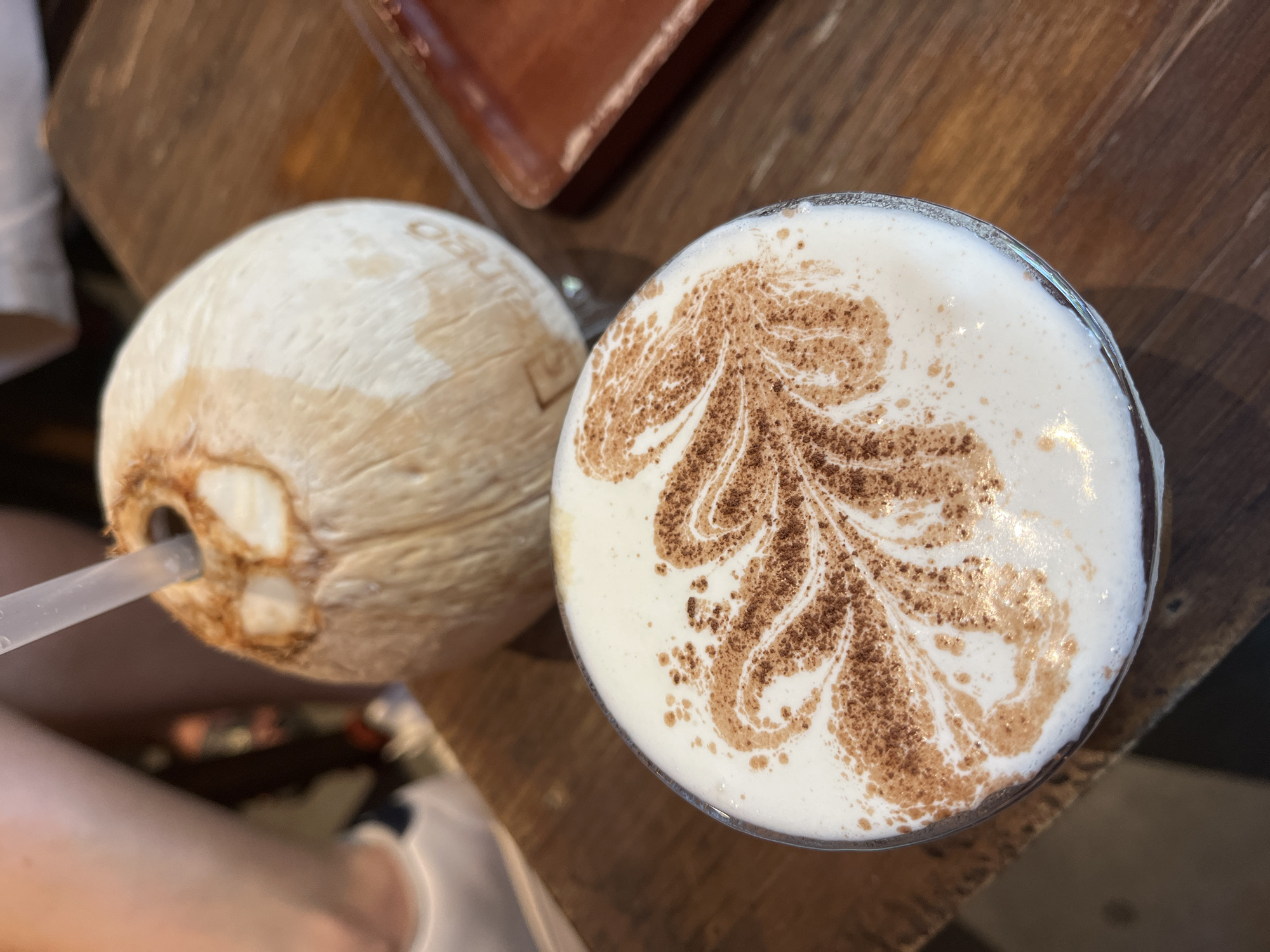 The scene depicts two drinks on a wooden table. One is a young coconut with a straw inserted. The other is a coffee beverage in a glass, topped with foamed milk decorated with a brown, leafy pattern. The location is Đà Nẵng, Vietnam. A person is partially visible in the lower left corner. A brown object rests partially out of focus on the table. The setting suggests a casual dining or café environment.