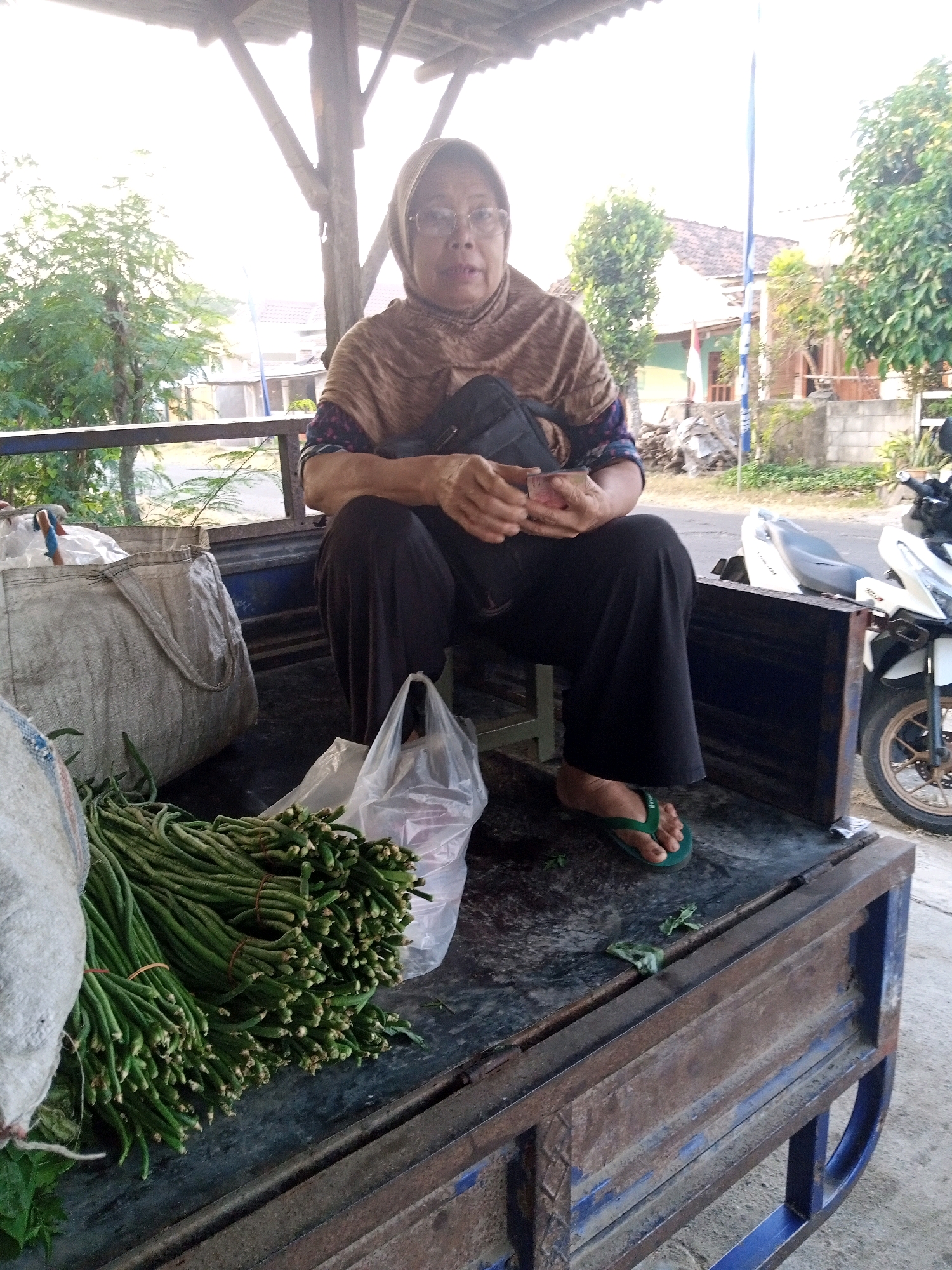 A woman wearing a brown hijab and glasses is seated on the back of a blue and rusted metal truck bed. She is wearing a brown patterned top and dark pants, with green flip-flops. She holds a small stack of bills in her hands and appears to be counting them. To her left is a large, off-white woven bag and a bundle of long green beans. To her right, a clear plastic bag holds more vegetables. A white motorcycle is parked behind the truck bed. The scene appears to be outdoors, possibly in a market or roadside stall, with trees and buildings visible in the background, suggesting a rural or suburban setting in Mojokendil, Indonesia.