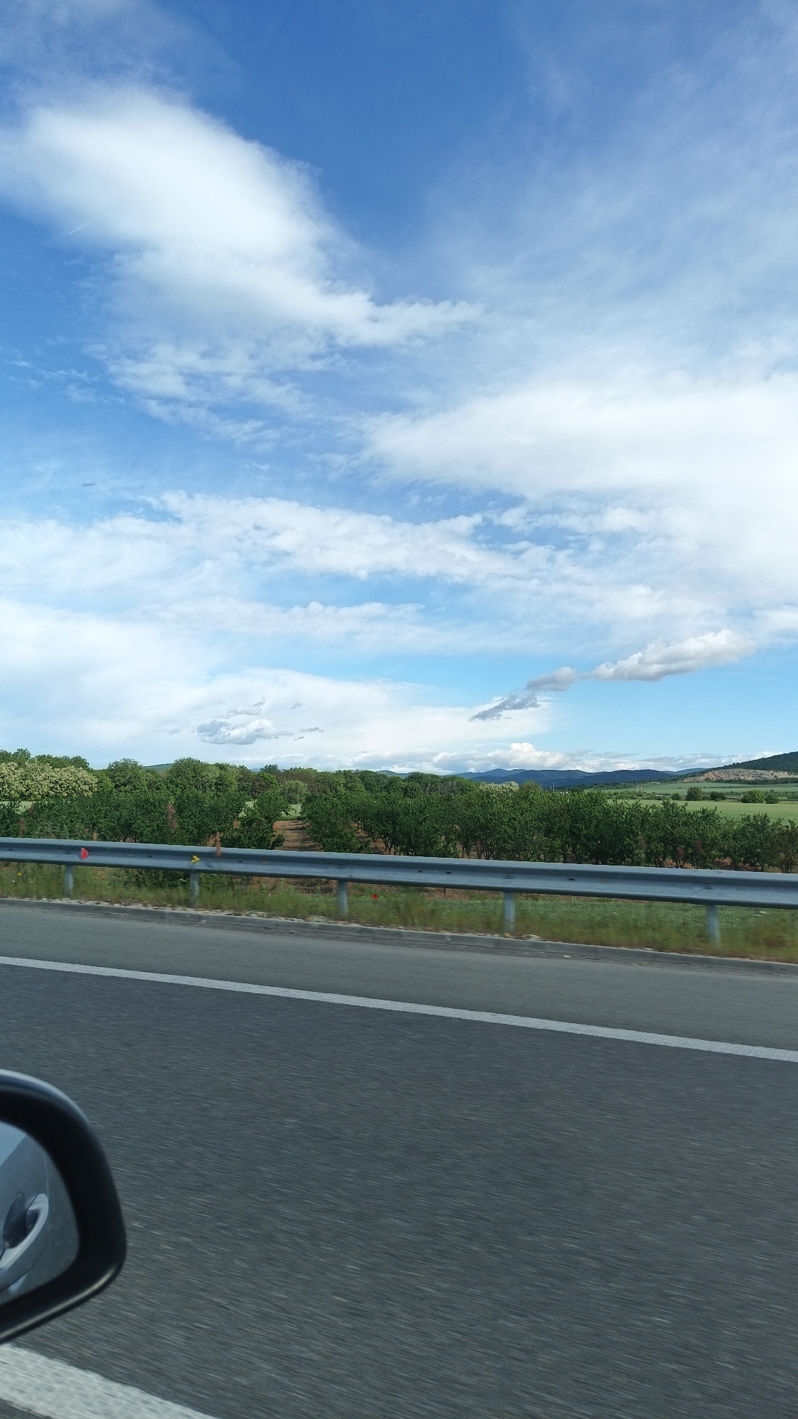 A view from inside a moving vehicle, likely a car, shows a multi-lane asphalt road with a white dividing line. A metal guardrail separates the road from a field with green trees and vegetation. In the background, rolling hills meet a sky filled with white and gray clouds against a blue backdrop. Part of the car's side mirror is visible in the lower left corner. The image captures a moment on a road, likely in a rural or semi-rural area of Bulgaria, possibly in or near Arnautito, given the provided location.