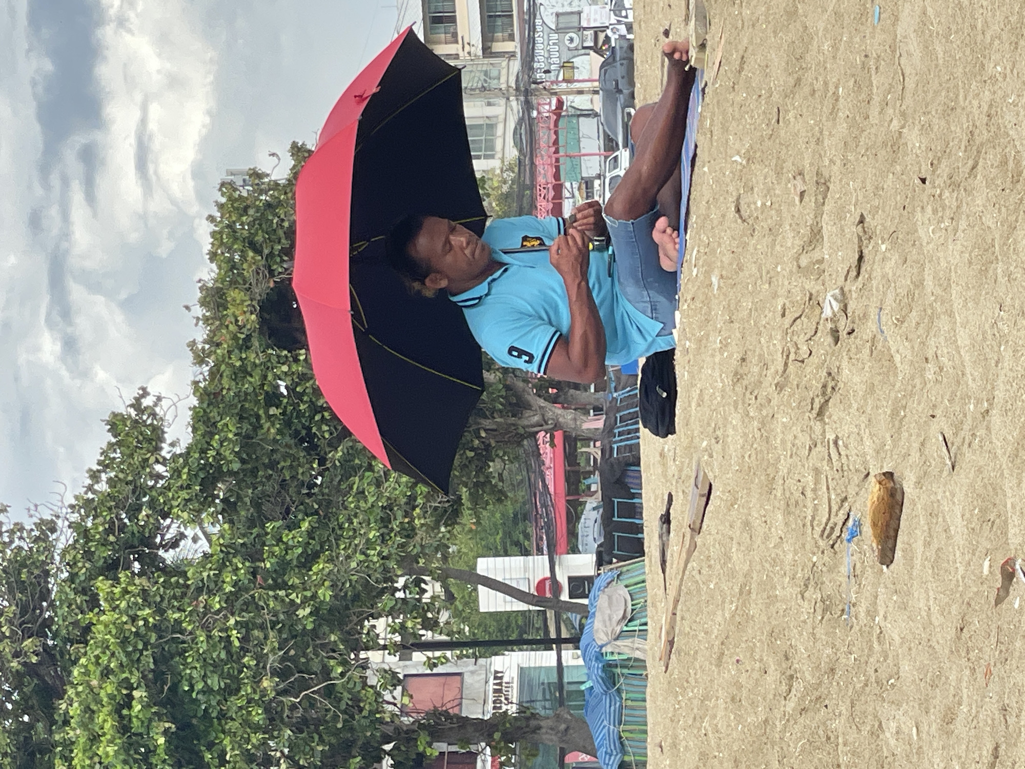 A man is sitting on a sandy beach in Pattaya City, Thailand, under a red and black umbrella. He is wearing a blue polo shirt with the number "9" on it and denim shorts. The man is holding something small in his hands and appears to be focused on it. The beach is littered with small pieces of debris. In the background, there are trees, buildings, and parked cars, suggesting an urban coastal environment. The sky is overcast.