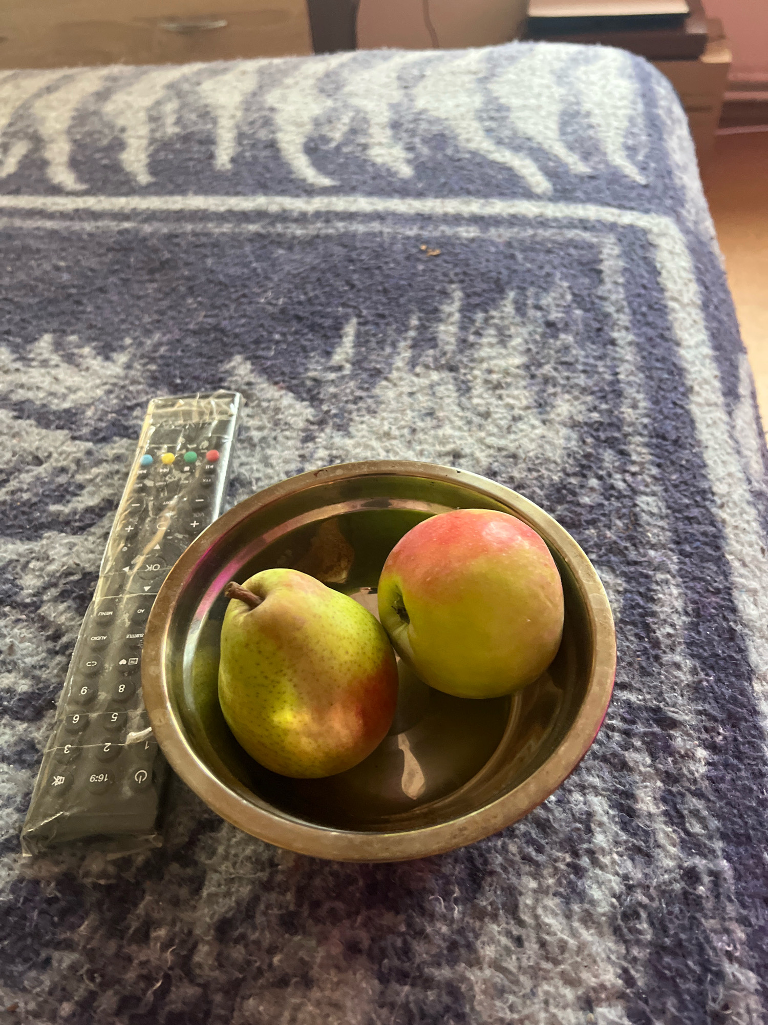 An overhead shot shows a silver bowl containing a pear and an apple, resting on a blue blanket with a white geometric pattern. To the left of the bowl, a black TV remote, partially covered in plastic, is placed on the blanket. The scene is lit by natural light coming from the right, casting soft shadows. The background is out of focus, showing furniture. The image was taken in Drohobych, Ukraine.