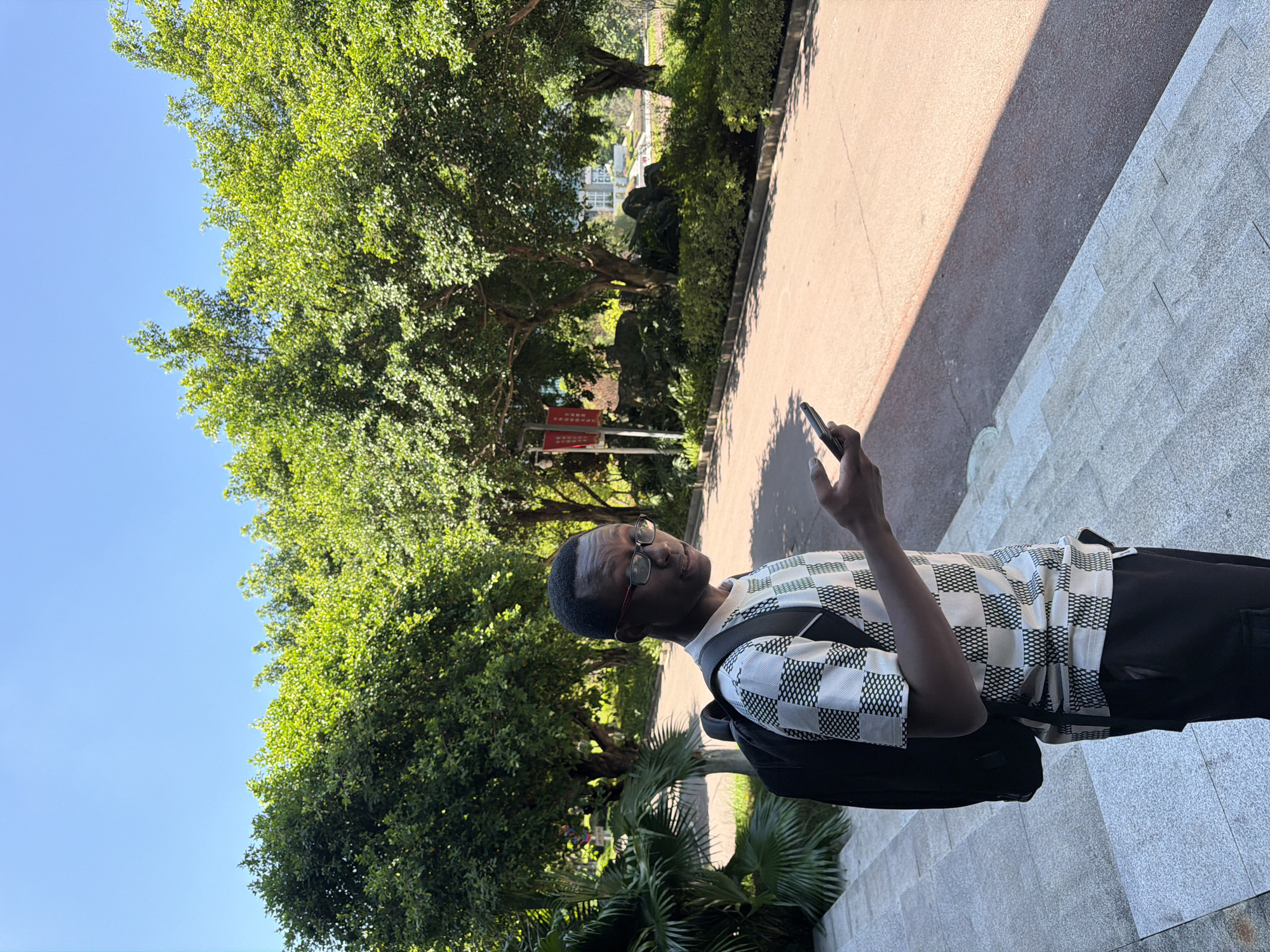 In a sunlit outdoor scene, a young Black man wearing glasses, a checkered shirt, and a backpack stands on stone steps, looking at his phone. The background features lush green trees lining a paved road, with a building visible in the distance. The image appears to be taken in the city of Jiangjin, China, a district of Chongqing, known for its scenic beauty and modern infrastructure.
