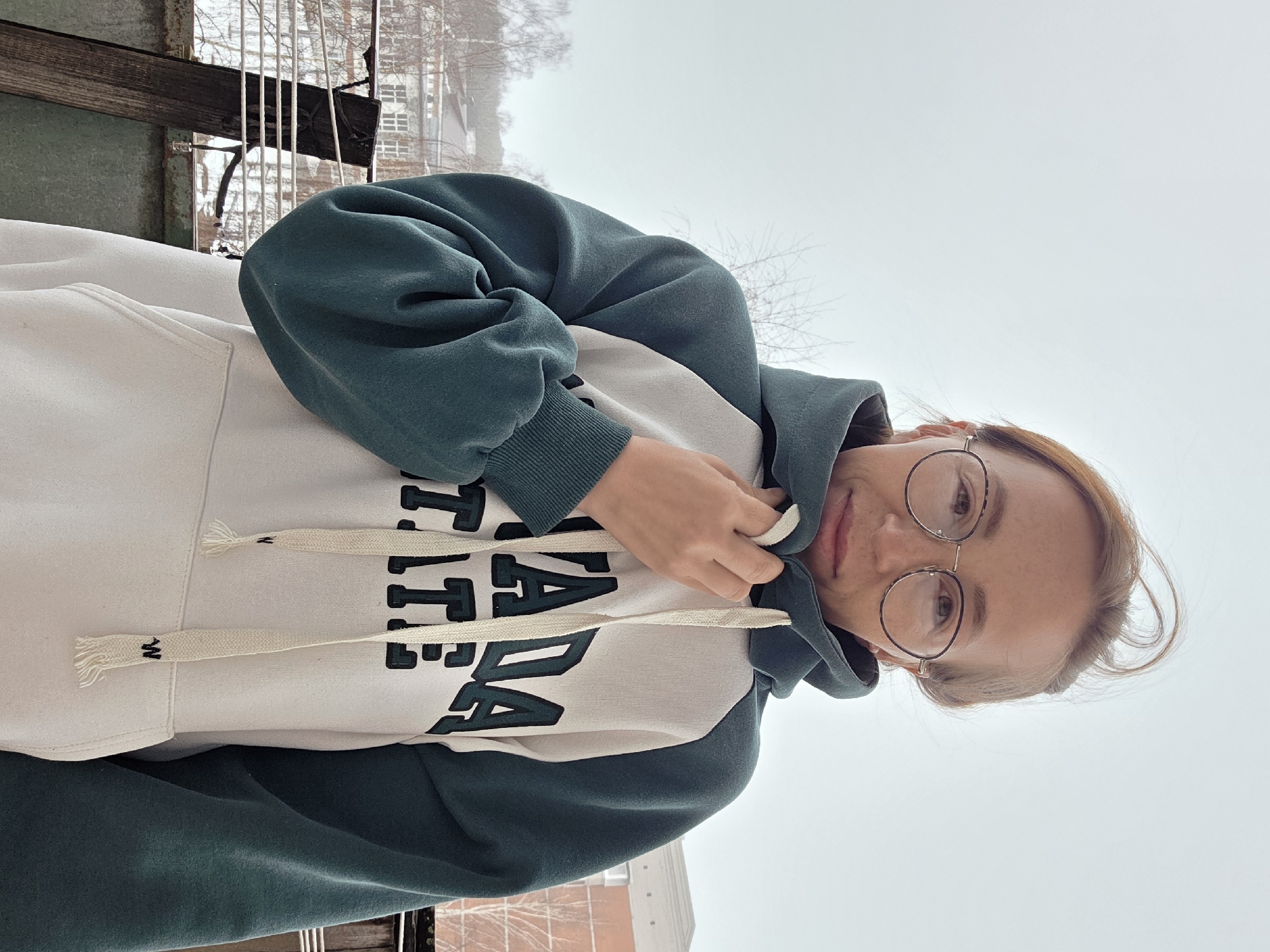 A young adult female is the primary subject, facing the camera while partially obscured by the hood of a cream-colored hoodie with dark green accents. The hoodie has the word "CANADA" printed on it in bold, black letters. She is wearing round-framed glasses. The background features an overcast sky and a blurred cityscape, including buildings and bare trees suggesting a cold season. Architectural details, such as window frames and building facades, are visible through the haze. The image was taken outside in Chaikovsky, Russia, as indicated by the visible cityscape elements.