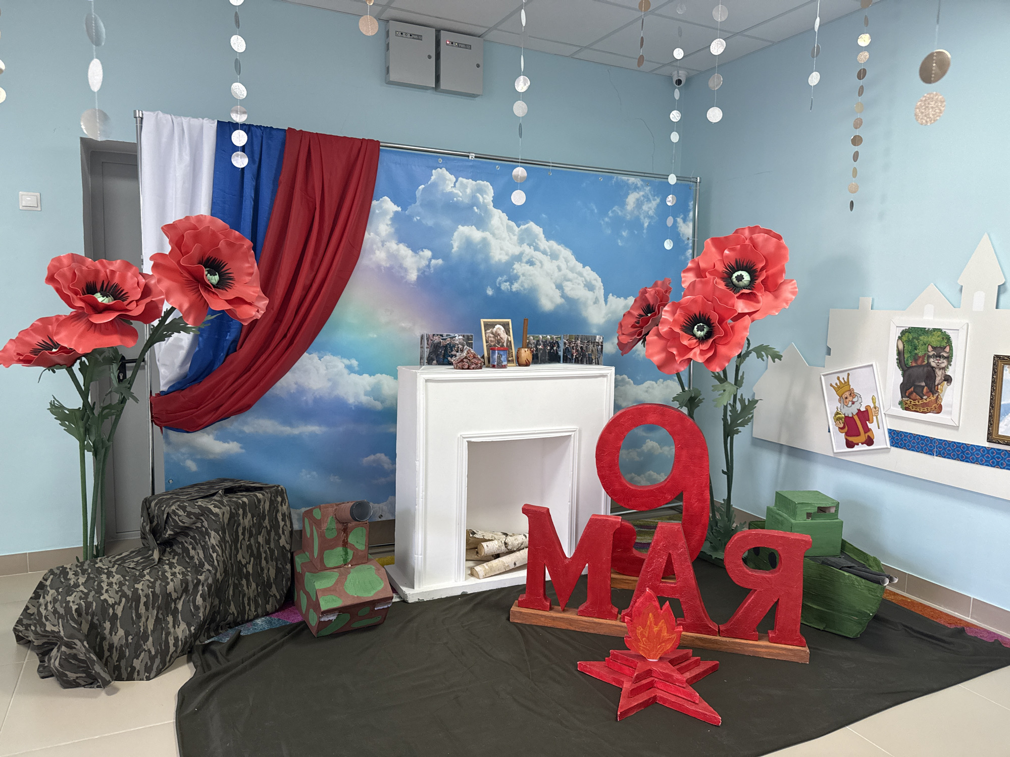 The image shows a room in Tobolsk, Russia, decorated for Victory Day, observed on May 9th. The backdrop consists of a white, blue, and red Russian flag draped against a background of blue skies with clouds. In front of the backdrop, there's a white faux fireplace with birch logs inside. Large red letters "9 МАЯ" (May 9th) are prominently displayed, along with a red star with a flame. The display also includes camouflage-covered objects resembling military equipment, and large red poppy flowers. Photographs are displayed on top of the faux fireplace. Silver garlands hang from the ceiling. Several illustrations are tacked to the wall on the right.