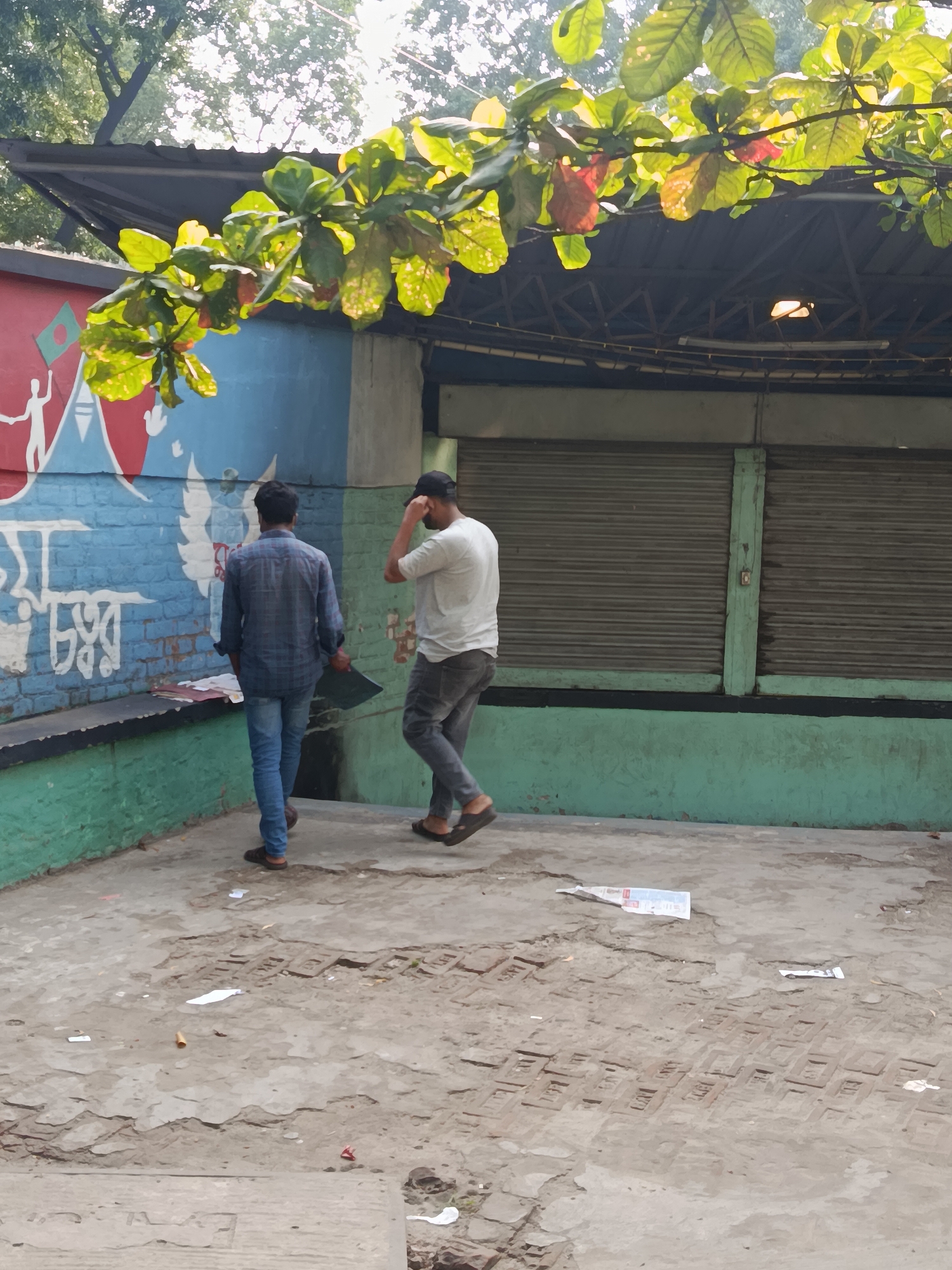 Two men are walking on a paved area covered with debris. The first man is wearing a blue shirt and jeans, and he is carrying a black folder. The second man is wearing a white shirt and gray jeans. They are walking towards a building with a mural on the left side and closed gray shutters on the right. The location is the city of Dhaka, Bangladesh. A tree branch with green leaves is hanging above them.