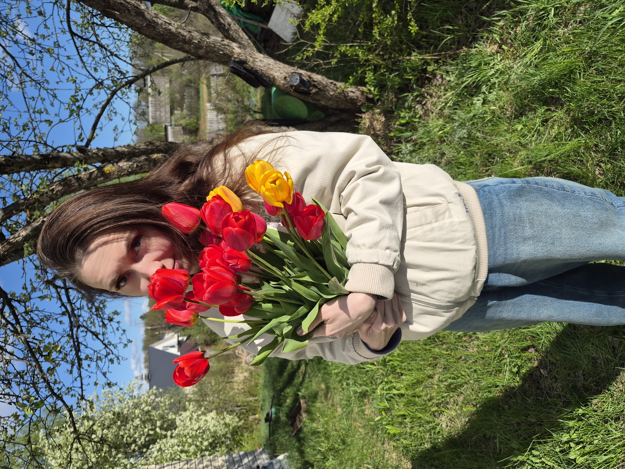 A woman with long brown hair holds a bouquet of red and yellow tulips. She is wearing a light-colored jacket and blue jeans. The background shows green grass, trees, a blue sky with some clouds, and houses. The location is Chaikovsky, Russia.