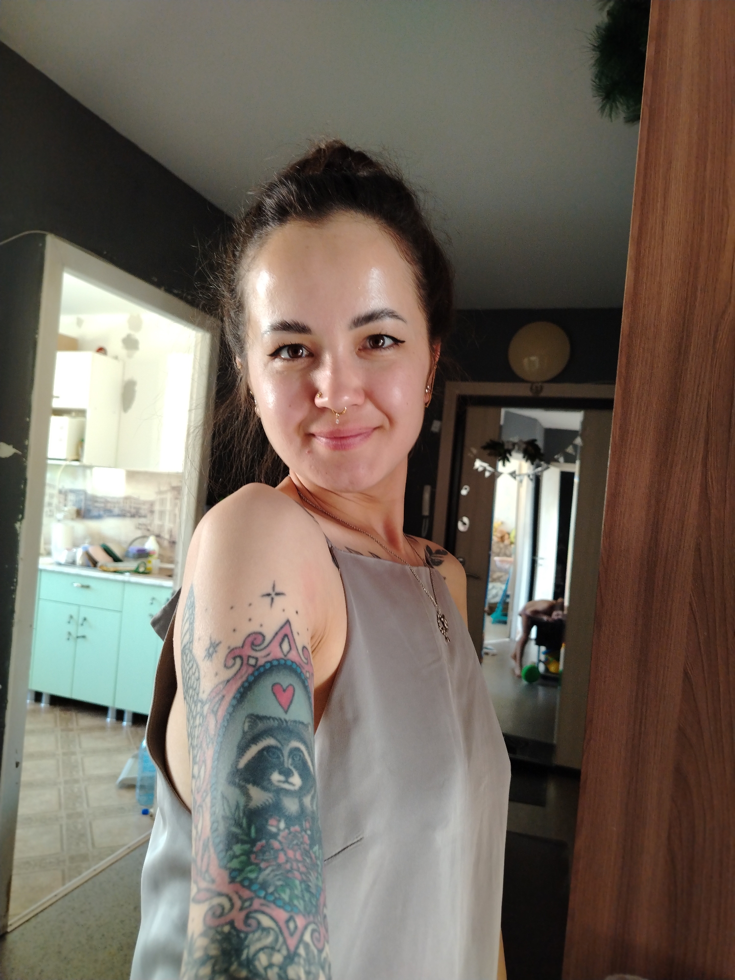 A woman with her hair in a bun stands in what appears to be an apartment. She is wearing a gray sleeveless top and has a tattoo of a raccoon framed by a pink heart on her upper left arm. She also has a nose piercing. A kitchen with light blue cabinets can be seen through a doorway behind her, and another room with a person bending over is visible through another doorway to the right. A wooden door stands partially open to the right. The location is likely Noviy, Russia, based on the user request.