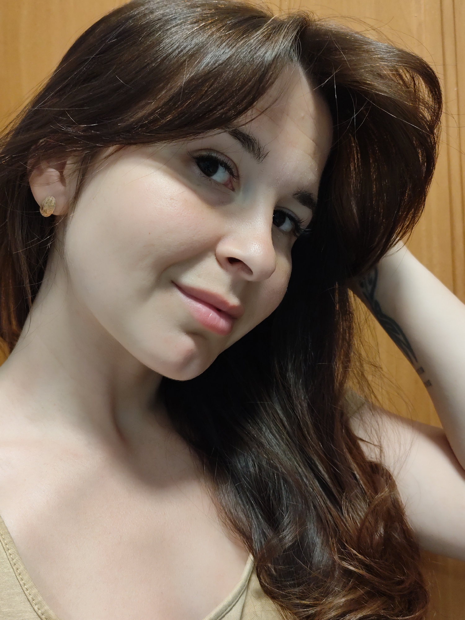 A woman is captured in a close-up, angled shot. Her brunette hair is styled with loose waves. She is wearing a tan shirt and a gold, circular earring. A tattoo is visible on her arm. The background is a wood-grain surface. The location is not determinable.