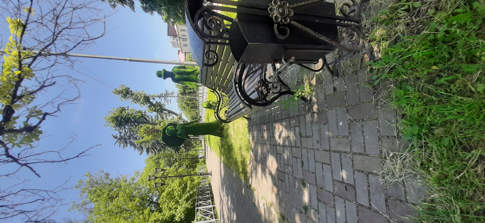 The image depicts a park scene in Yessentukskaya, Russia, under a clear blue sky. In the foreground, a dark metal park bench with a decorative design is positioned on a brick-paved pathway. To the right of the bench is a black metal trash receptacle. Behind the bench and to the left, a paved pathway curves through a grassy area. The pathway is lined with a white metal fence. Several lampposts are visible along the pathway. The main subjects of the scene are two topiary figures, shaped like people. The figure on the left appears to be a woman holding an umbrella. The figure on the right is dressed in a hat and coat. A smaller topiary figure, possibly an animal, is located between them. Lush green trees and foliage surround the area, indicating a warm season. The lighting suggests it is daytime.