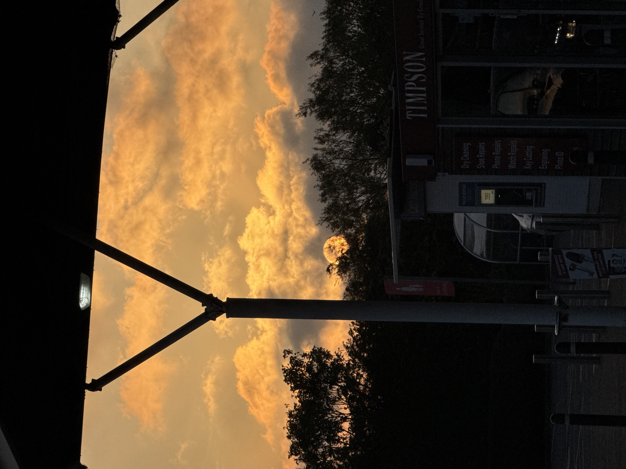 The image shows a sunset sky with orange-tinted clouds. The sun is partially obscured by trees. In the foreground, there is a dark structure with a light fixture. A Timpson shop is visible, offering dry cleaning, shoe repairs, phone repairs, watch repairs, key cutting, engraving, and photo ID services. There's also a Tesco self-service point. A poster advertises household and car keys and passport/visa photos. The shop's sign reads "TIMPSON Great Service By Great People". The location is Great Yarmouth, United Kingdom. The image is oriented sideways.