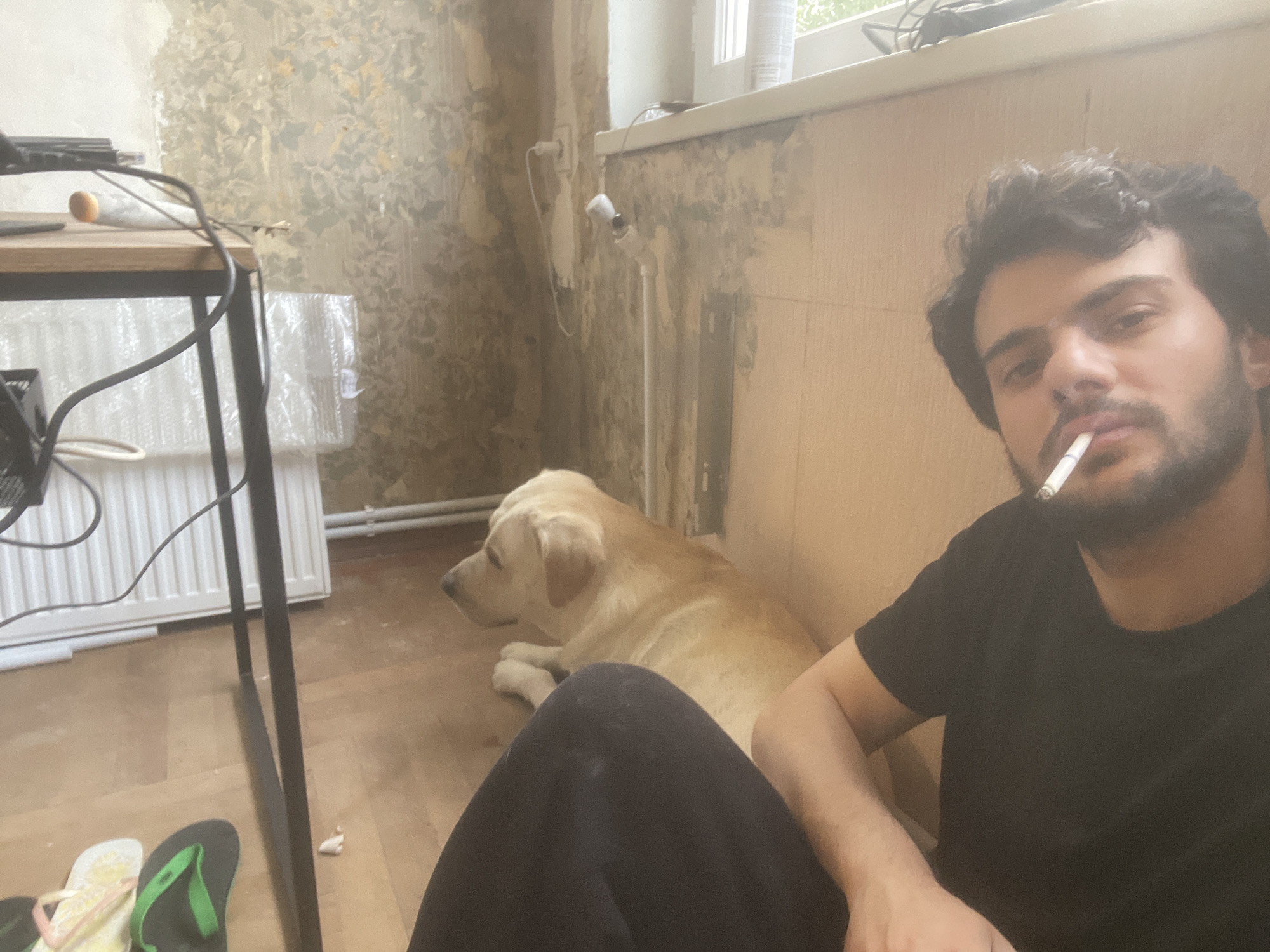 A man with dark hair and a beard is sitting on the floor, smoking a cigarette. He is wearing a black t-shirt and dark pants. Beside him lies a light-colored Labrador retriever. In the background, a wooden table with some electronics and wires is visible, along with a radiator and a window with a view of what appears to be an urban setting, possibly Tbilisi, Georgia, indicated by the style of the building and the weathered wallpaper. The room has a rustic and somewhat neglected appearance.