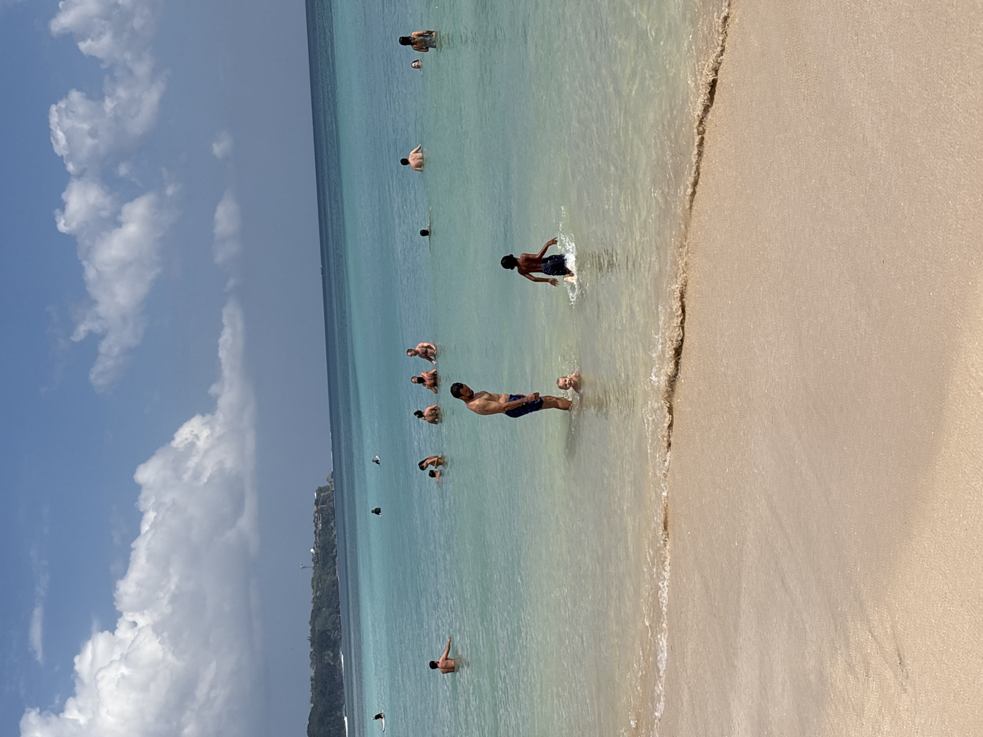 A daytime aerial shot shows a beach scene in Kuta Selatan, Indonesia, with the ocean and shore in clear view. The water is a vibrant turquoise, gradually deepening to a darker blue further out. A gentle wave laps at the tan, sandy shore. Several people are visible in the water, some swimming, others standing or wading. In the foreground, a man and a young child are in shallow water. A group of people are scattered further out, with some appearing to be socializing. The distant coastline features lush greenery and structures, suggesting a developed area. The sky is a bright blue, dotted with fluffy white clouds.