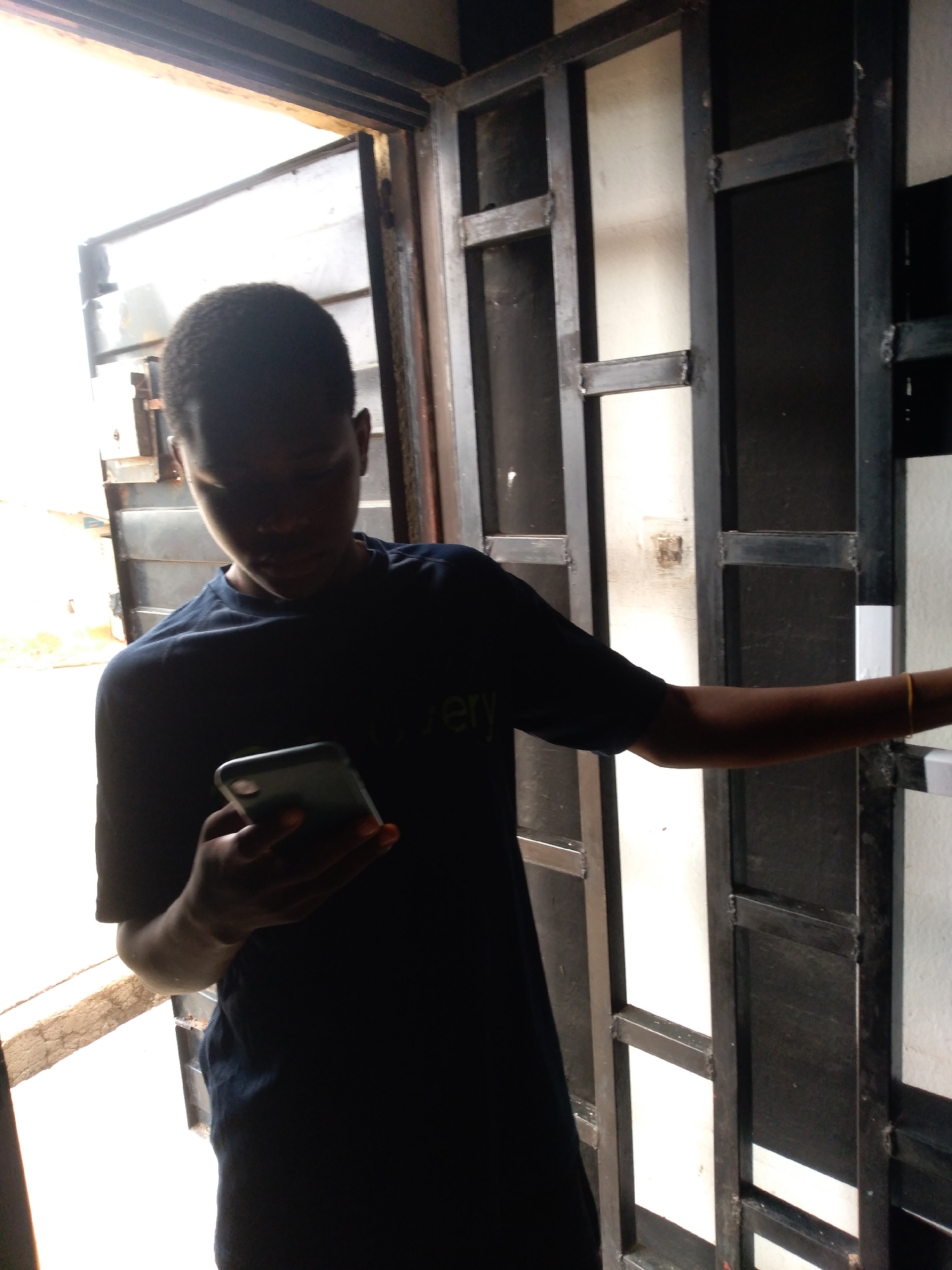 A young boy, appearing to be in his early teens, stands at an open doorway, illuminated by bright natural light. He is wearing a dark blue t-shirt and holding a smartphone in his right hand, looking down at the screen. The boy's left arm is extended, resting on a black metal frame that appears to be part of a gate or door. He has short, dark, curly hair. The background consists of a black metal grid structure, likely a gate, with white panels visible behind it. The setting suggests an outdoor or semi-outdoor location, possibly a residential entrance.