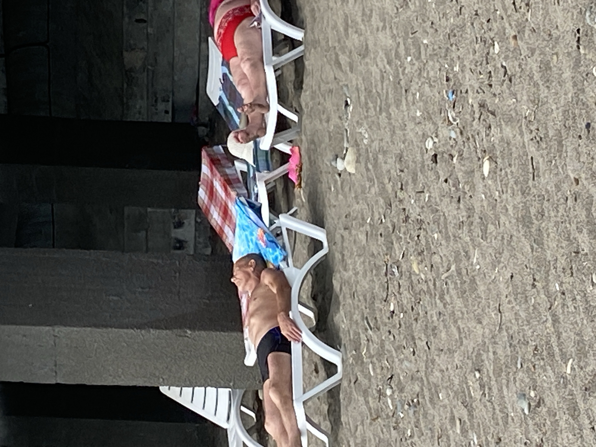 Two individuals are sunbathing on beach chairs on a sandy beach. The person on the left is a male wearing black swim trunks, lying on his back with his head tilted back. The person on the right is a female wearing red bikini bottoms, lying on her back with her legs extended. Beach chairs, towels, and a hat are visible. The background shows large concrete pillars and a dark overhang, suggesting a sheltered area or structure. The beach appears to be located in Sokhumi, Abkhazia.