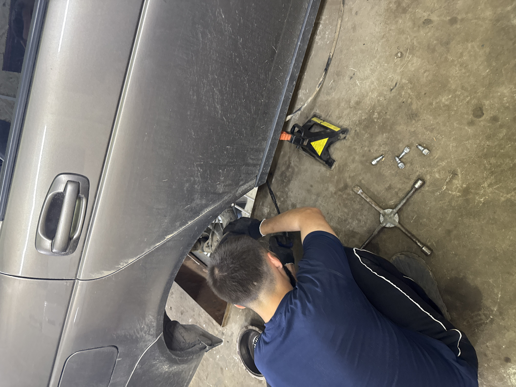 This image, depicted in Perm, Russia, captures an automotive repair scene. A male individual, observed from behind, is kneeling on a concrete floor while working on the rear section of a metallic grey sedan. He is wearing a dark blue short-sleeved t-shirt and black track pants with white side stripes, along with black gloves. His head is bent down as he attends to components within the exposed rear wheel well, where the wheel is absent.
The car, heavily soiled with dirt and mud splashes across its lower panels and fender arches, is elevated and supported by a yellow and black jack stand positioned under its right rear rocker panel. The fuel filler cap is visible on the rear quarter panel. On the concrete floor, a cross-shaped lug wrench rests, alongside four silver-colored lug nuts scattered nearby. To the left of the kneeling individual, a round object, possibly a brake drum, is also on the floor. The ground itself is a dirty, stained concrete surface, consistent with a workshop or garage environment. In the background, under the car, a wooden beam or similar structure is visible.