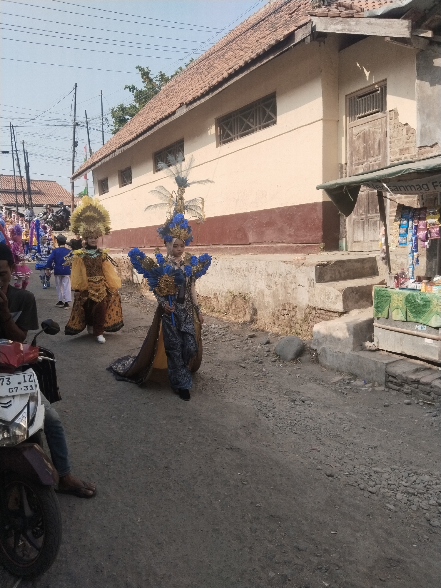 In the city of Jembayat, Indonesia, on a dirt road, two young women dressed in elaborate, colorful costumes are participating in a parade. The woman on the left wears a yellow and brown patterned outfit adorned with a tall, feathered headdress. The woman on the right is in a blue and gold costume with a large, feathered collar and a long train. They are walking down the road, which is lined with buildings. In the background, other parade participants and spectators can be seen. A motorcycle is parked on the left side of the road, in the foreground. The scene suggests a festive cultural event taking place.