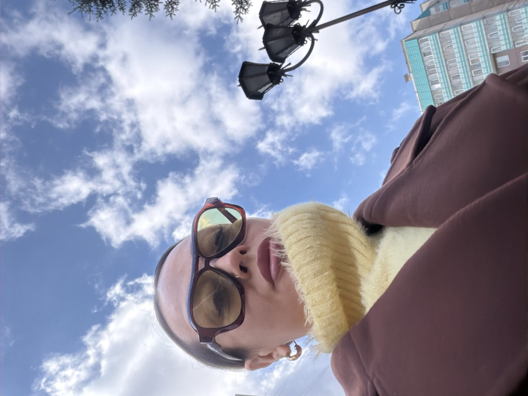 A low-angle, close-up shot captures a person outdoors against a bright blue, partly cloudy sky. The individual, who appears to be female, is wearing large, dark-rimmed sunglasses with reflective lenses that obscure their eyes. Their lips are slightly parted, and a subtle nose piercing is visible. They are dressed in a light yellow, high-necked or turtleneck sweater with a ribbed texture, layered under a dark brown or maroon jacket or coat. A gold-colored hoop earring is seen on their visible right ear.
The perspective is looking upwards, with the sky dominating the upper portion of the frame. In the upper left corner, dark green coniferous tree branches are partially visible. Towards the upper right, an ornate street lamp with multiple fixtures extends into the sky. Further in the background, a multi-story building with a distinctive facade featuring light blue and white/grey horizontal banding and numerous windows is visible, indicating an urban setting.
The image conveys a daytime scene with fair weather, suggesting coolness due to the person's attire. The location is an urban outdoor environment, which, combined with the clothing, is consistent with the provided context of Krasnoyarsk, Russia. There is no discernible text visible in the image.