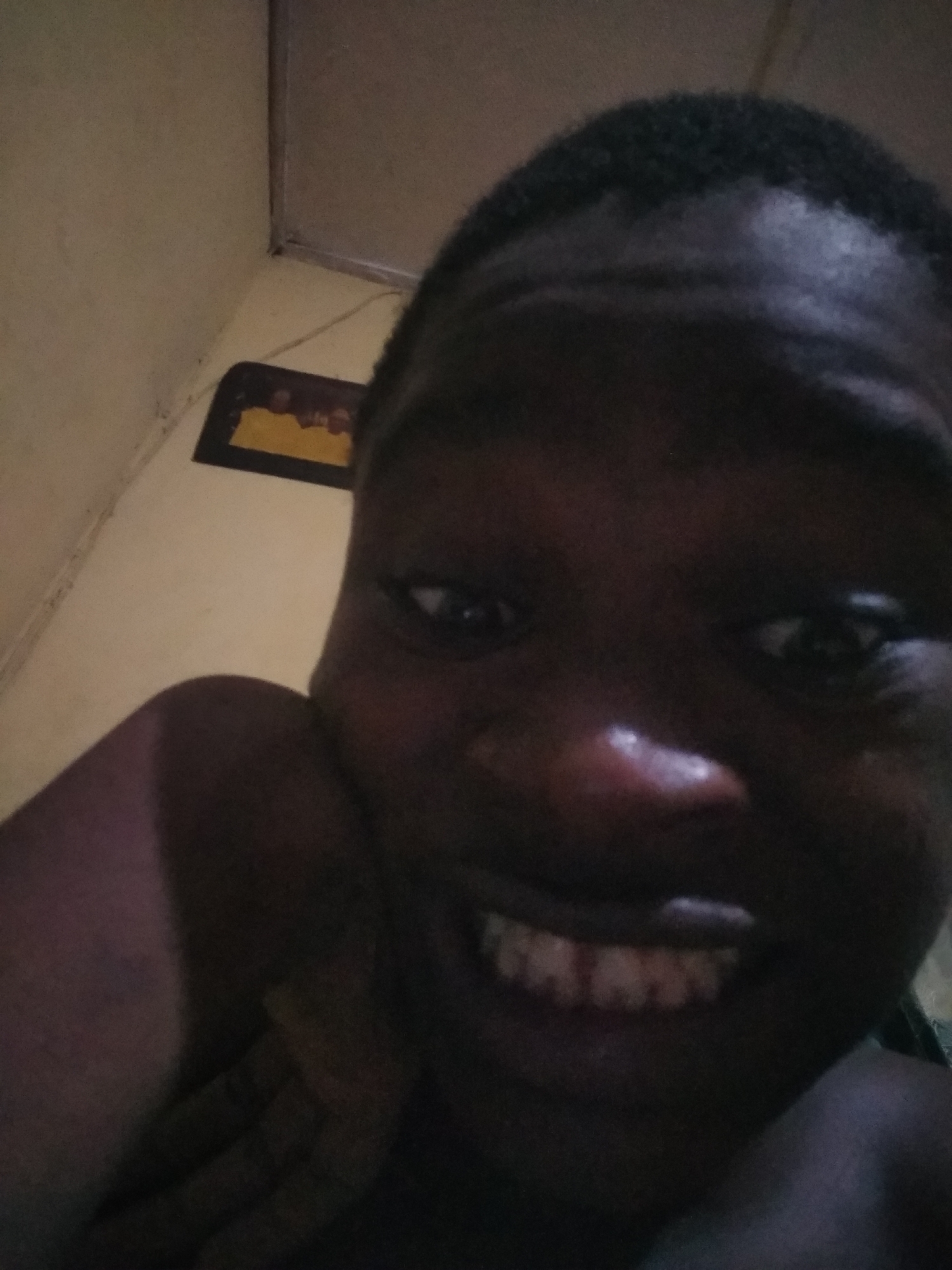 A close-up selfie of a dark-skinned individual with short black hair. The individual is smiling, revealing teeth. The background is a dimly lit room with a cream-colored wall. A picture frame with a yellow border hangs on the wall in the upper left corner. The setting is implied to be Shangisha, Nigeria, though this is not visually verifiable in the image. No specific actions or interactions are apparent beyond the act of taking the selfie.