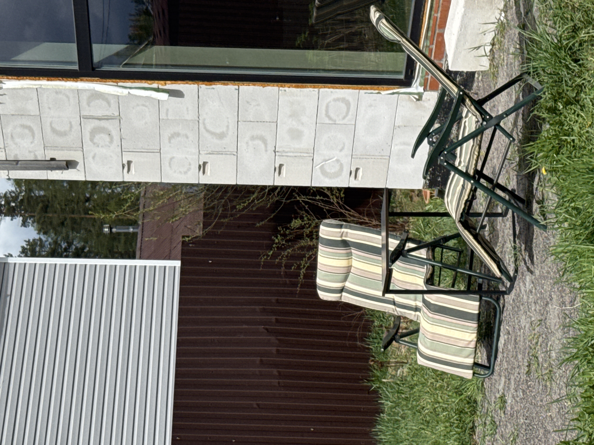 The image depicts an outdoor scene adjacent to a building, possibly in Steklyannyi, Russia. Two folding chairs with striped fabric are situated on a paved surface overgrown with grass. The chairs are positioned near the exterior wall of the building, which features a large window, light gray concrete blocks with circular stains, and both corrugated gray and brown siding. A small tree or shrub is visible in the background. No people are present.