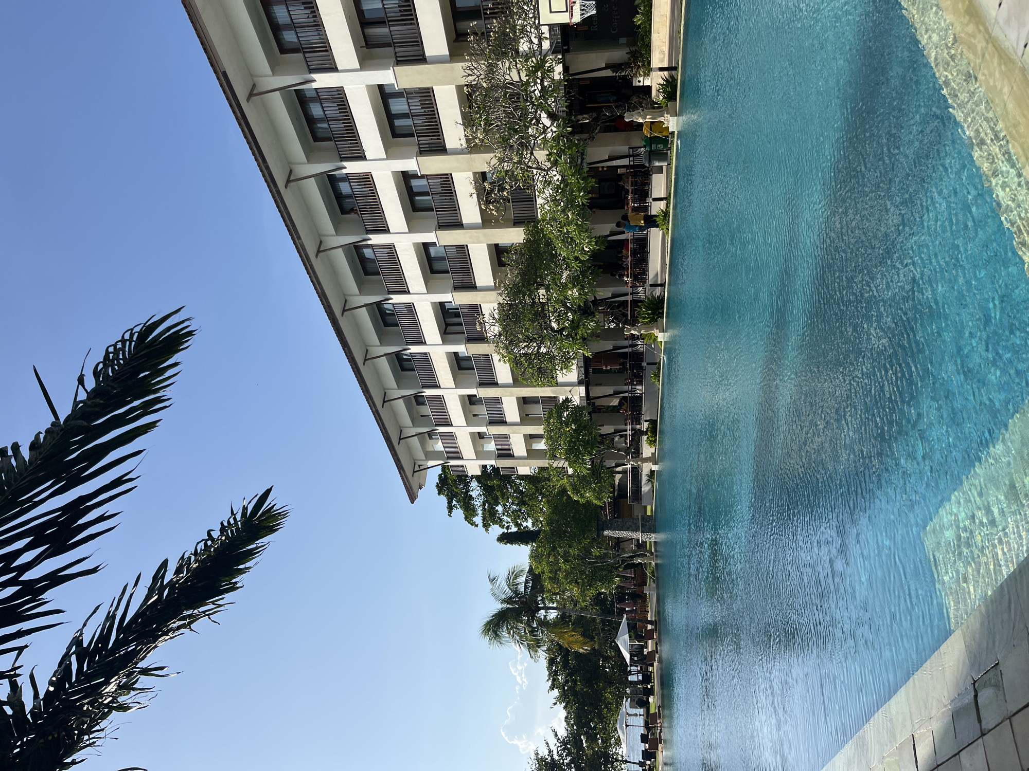 The image depicts a resort in Kuta Selatan, Indonesia, featuring a large, clear blue swimming pool in the foreground. Beyond the pool, a multi-story hotel building with balconies and dark-framed windows lines the back of the property. Lush green trees and palm trees are scattered around the hotel and pool area, with white umbrellas and lounge chairs visible near the water's edge in the distance. The sky is a clear, bright blue with a few scattered clouds. A portion of a palm frond is visible in the upper left corner, casting a shadow on the scene. Several people are present, some sitting at tables in the outdoor dining area of the hotel, while others are standing near the pool. The overall atmosphere is one of a tranquil and sunny resort environment.
