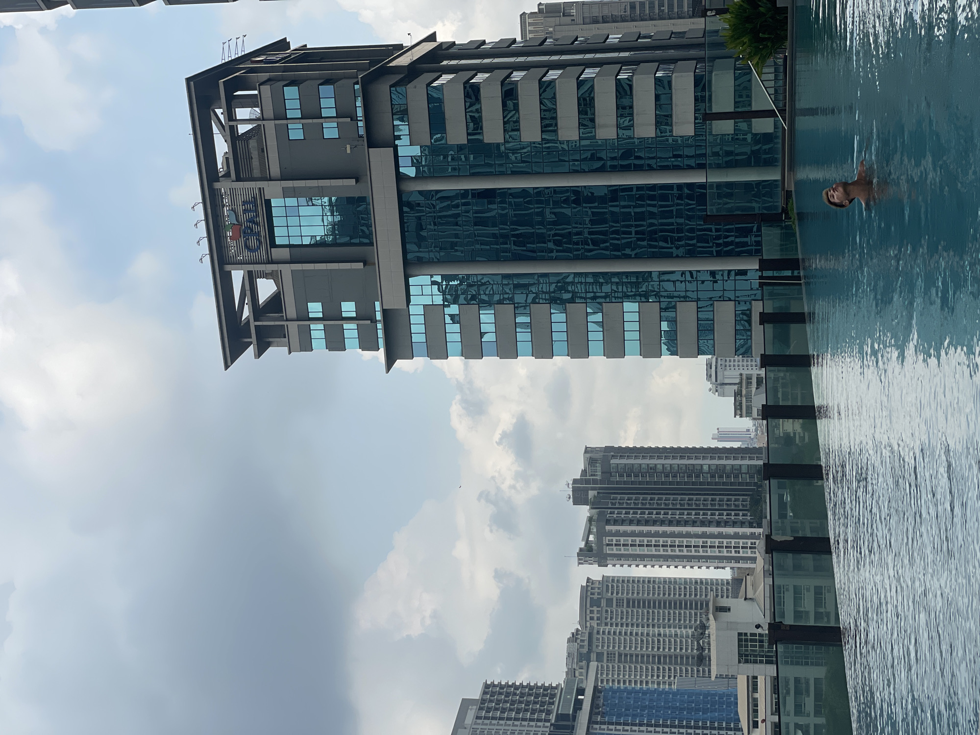 The image, correctly oriented, displays a vibrant urban scene featuring an infinity swimming pool overlooking a city skyline on a bright, clear day.
In the foreground, an infinity pool with rippling blue water occupies the right side of the frame. A person is partially visible in the pool, with their head and shoulders above the water, facing towards the left, appearing relaxed. A small palm tree is visible near the edge of the pool.
Dominating the left side of the image is a modern high-rise building with a sleek, grey facade and numerous blue-tinted glass windows. Reflections of the sky and surrounding environment are visible on its glassy surface. On one of the upper levels of this building, a multicolored sign with the text "CUBE" is clearly visible, with the letters appearing to be green, red, blue, and possibly yellow. Various architectural features and what appear to be antennas or equipment are also present on the building's top.
In the midground and background, an expansive urban landscape stretches out, filled with numerous other high-rise buildings and skyscrapers, typical of a bustling metropolis. The sky above is mostly clear with scattered white clouds, indicating sunny weather. The overall impression is one of leisure and urban luxury, likely a rooftop pool setting in a major city like Bangkok, Thailand, during the daytime.