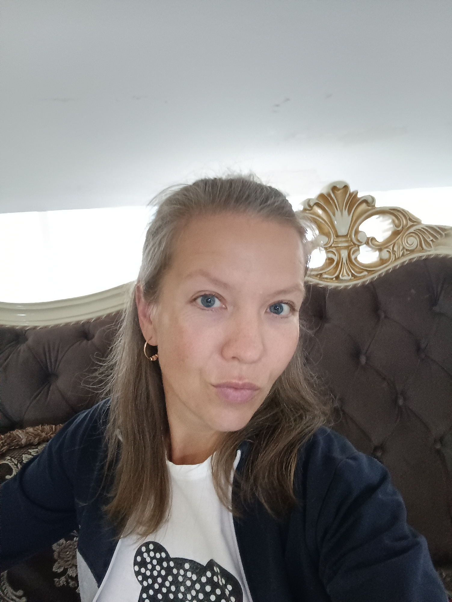 The image shows a medium shot selfie of a woman with blonde hair, blue eyes, and fair skin. She is wearing a white t-shirt with a black polka-dot bear design and a dark blue jacket. She is sitting on a brown tufted sofa with a gold ornate headrest. The background is a plain white wall. The woman's expression is neutral, with her lips slightly pursed. The image appears to have been taken indoors in a domestic setting. There is no specific identifiable location within Yekaterinburg, Russia, that can be determined from the image.