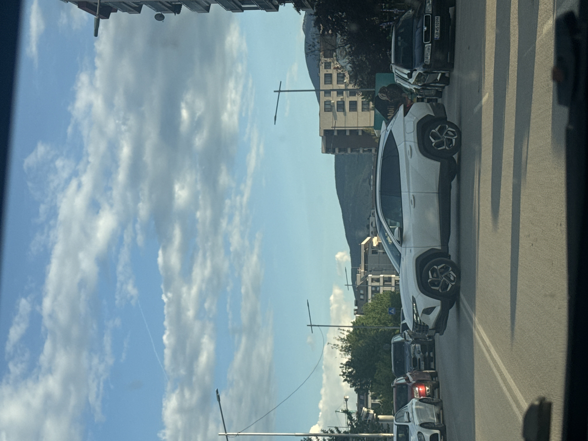 The image, taken from inside a vehicle, shows a street scene in Sofia, Bulgaria. In the foreground, a line of cars is visible on the right side of the road. The second car in line is a white crossover with distinctive wheels. Behind the white car is a dark-colored sedan, its license plate visible. The sky is partly cloudy, with scattered cumulus formations. In the background, a mountain range is discernible, partially obscured by buildings. Streetlights are scattered along the road.