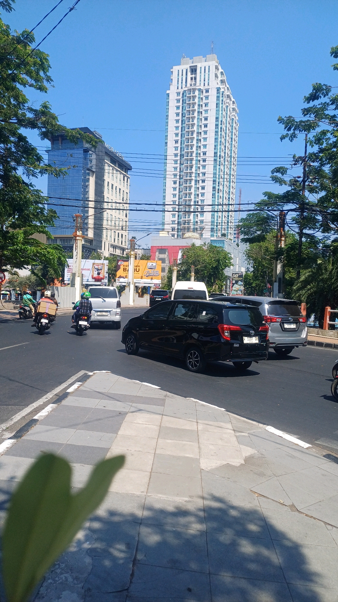 The image captures a daytime street scene in Surabaya, Indonesia, with several vehicles and buildings visible. Dominating the background is a tall, modern white skyscraper with numerous windows, characteristic of urban development. To its left stands another building with a darker facade and "MORAZEN" signage. The street is paved and features a pedestrian sidewalk with gray square tiles.
In the foreground, a black car is driving on the road, with a silver SUV behind it. To the left, two motorcycles are seen, one carrying a large brown box on the rear. The road is busy with traffic, and there are power lines overhead. Green trees line the sides of the street, casting shadows on the sidewalk. Billboards are visible in the mid-ground, indicating commercial activity. The sky is clear and blue. The overall impression is of a bustling city environment on a sunny day.