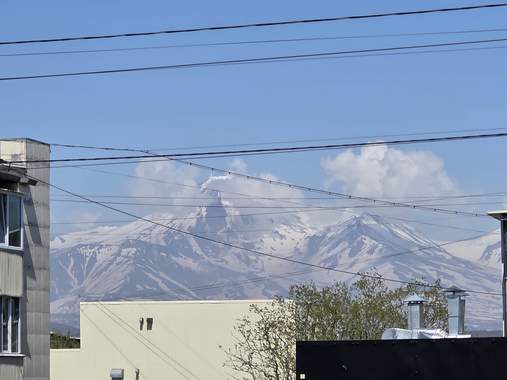 A medium shot captures a distant view of snow-capped mountains under a clear blue sky with scattered white clouds. In the foreground, power lines crisscross the frame. To the left, the corner of a building with vertical ribbed siding and windows is visible. A lighter-colored building with a flat roof occupies the mid-ground, with a portion of a dark structure in the lower right corner. Trees with sparse foliage are interspersed between the buildings and the mountains. Two metal ventilation pipes with conical tops are prominent on the roof of the lighter-colored building.