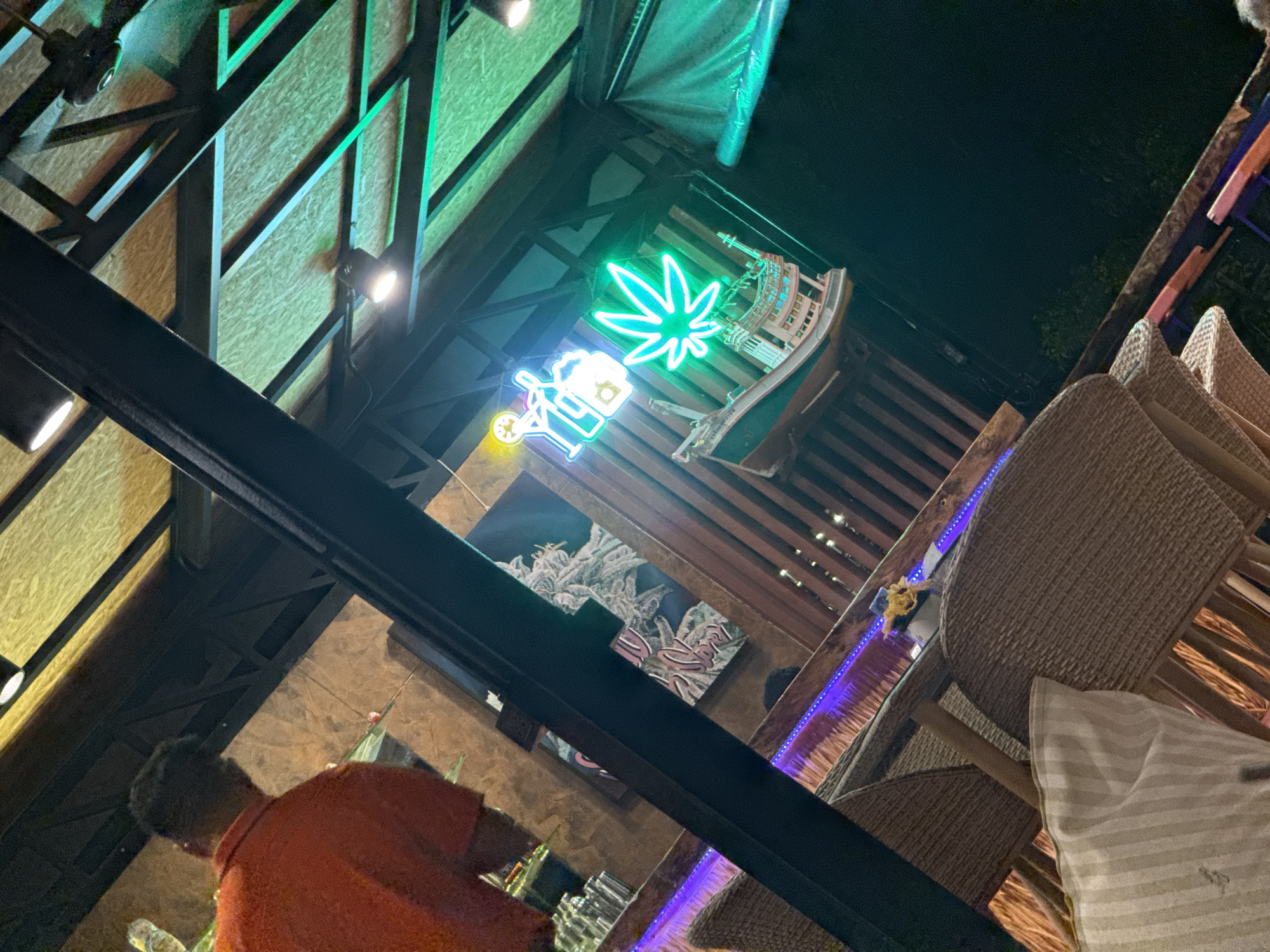 The image depicts an outdoor seating area, likely a restaurant or bar, in Ko Samui District, Thailand. The setting is dimly lit, with neon lights casting a vibrant green glow on the scene. A person wearing an orange shirt is visible in the background, their back to the camera. In the mid-ground, several woven chairs surround tables. Above the seating area, a model boat hangs suspended, adorned with neon lights forming a marijuana leaf and a beer mug with a figure. The ceiling structure is made of wood with visible beams and lights. The overall impression is of a casual, possibly entertainment-focused venue.