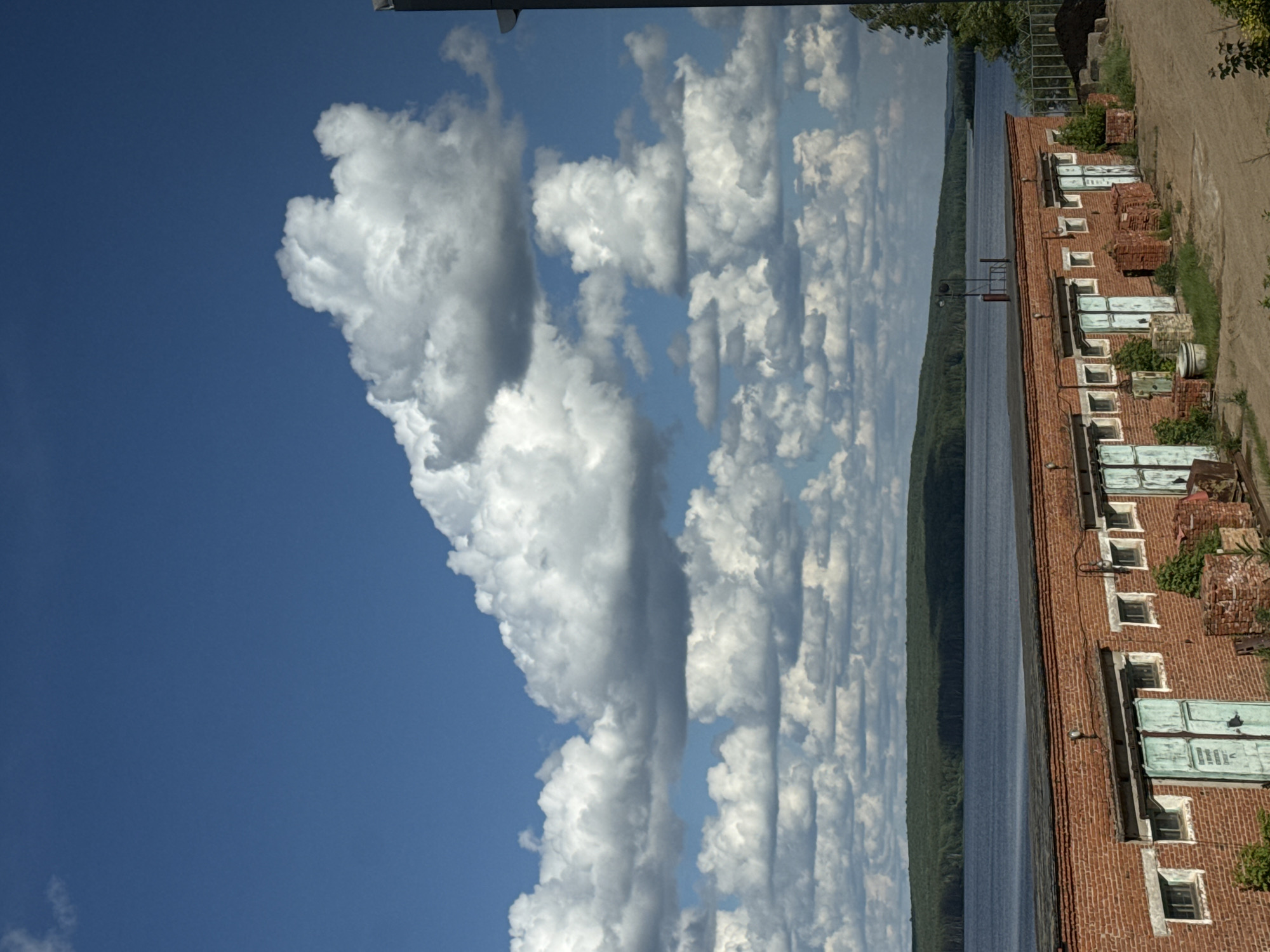 The image shows a view in Chaikovsky, Russia, featuring a body of water, likely the Kama River, with a forested shoreline visible in the background. Adjacent to the river is a long, low brick building with regularly spaced windows, some with green shutters or coverings. The building appears to be old and possibly abandoned or in disrepair. Above, a blue sky is filled with numerous cumulus clouds, with a large, prominent cloud formation to the left. A dirt path runs along the right side of the building. The overall scene suggests a tranquil setting with elements of nature and decaying infrastructure.
