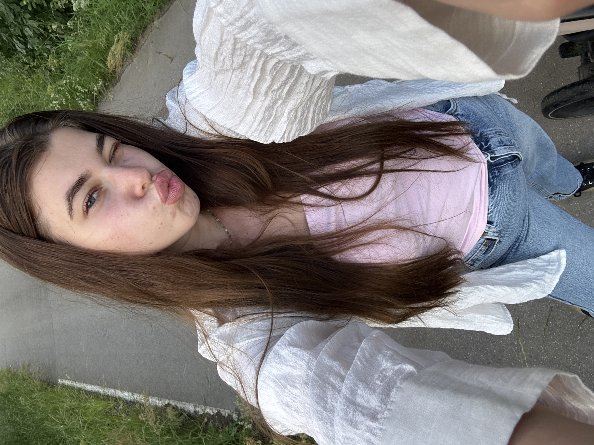 A young woman with long, brown hair is taking a selfie outdoors. She is winking and pouting her lips. She is wearing a pink crop top, blue jeans, and an open white shirt. She has a necklace with colorful beads. The background is a paved path with grass and trees on either side, suggesting a park or a roadside. The woman appears to be in motion, possibly riding a bicycle or scooter given the blurred wheel visible at the bottom of the frame. The scene is brightly lit, indicating it is daytime. There is no explicit visual information within the image that allows for precise identification of the location as Мещёры, Russia.