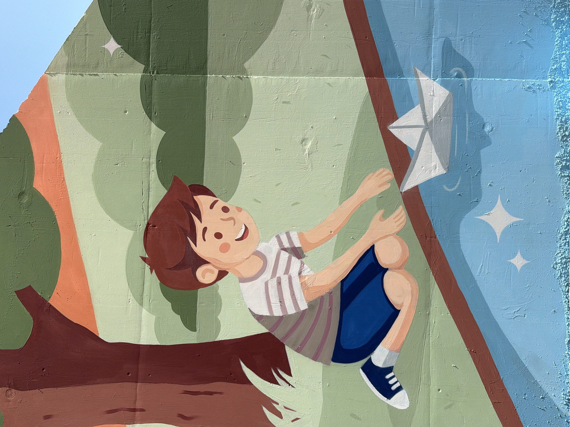 This is a mural painted on a wall, depicting a young boy kneeling by a body of water, about to release a paper boat. The boy has brown hair and is wearing a striped t-shirt and blue shorts. The water is a vibrant blue, and the paper boat is white, casting a shadow beneath it. In the background, there are stylized trees and green landscape elements, with a bright blue sky at the top. The mural has a textured surface, characteristic of being painted on a brick wall. The style is illustrative and cartoon-like. The exact location in Yekaterinburg, Russia, is not discernible from the image itself.