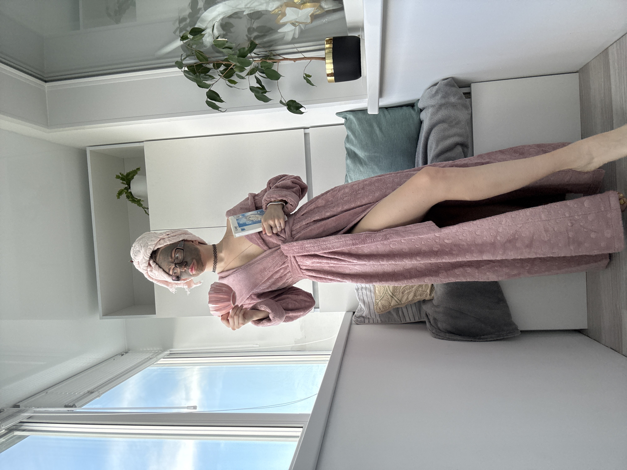 A person with a pink towel wrapped around their head and face mask is standing on a balcony and holding a pink cup and a book. They are wearing a pink robe and a necklace. The balcony has a window with blinds and a view of the sky. There is also a plant and some cushions in the scene.