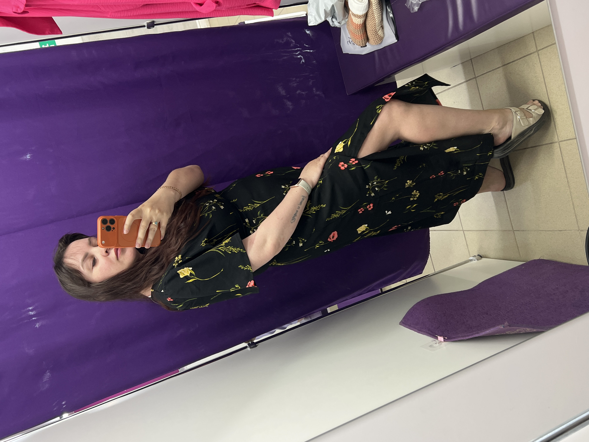 A woman lies on a purple surface in what appears to be a fitting room. She holds an orange smartphone and is taking a selfie. The background features a padded purple wall and a bench with shoes and bags on it. The floor is tiled. A purple cushion is also visible. There is no evidence to pinpoint this image's location to Noviy, Russia.