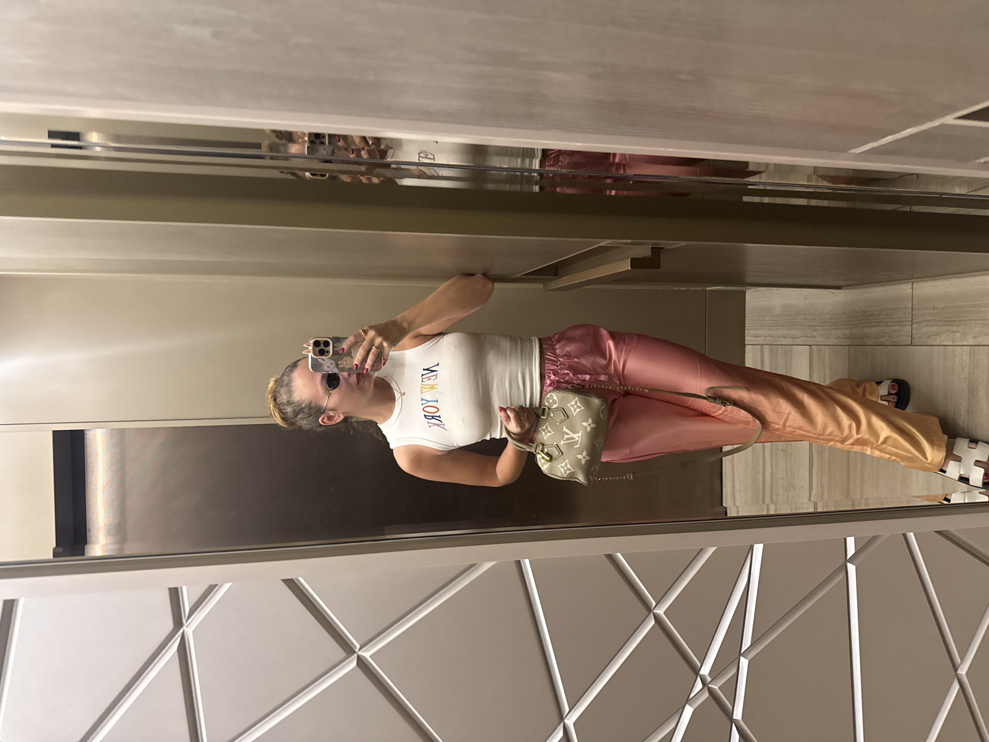A woman is taking a selfie in a mirror, possibly in an elevator or a similar enclosed space with reflective surfaces. She is wearing a white tank top with "NEW YORK" written on it, pink satin pants, and sandals. She is holding a handbag. The walls are decorated with geometric patterns. The location is uncertain; the city of Khimki, Russia is not identifiable within the visible context.