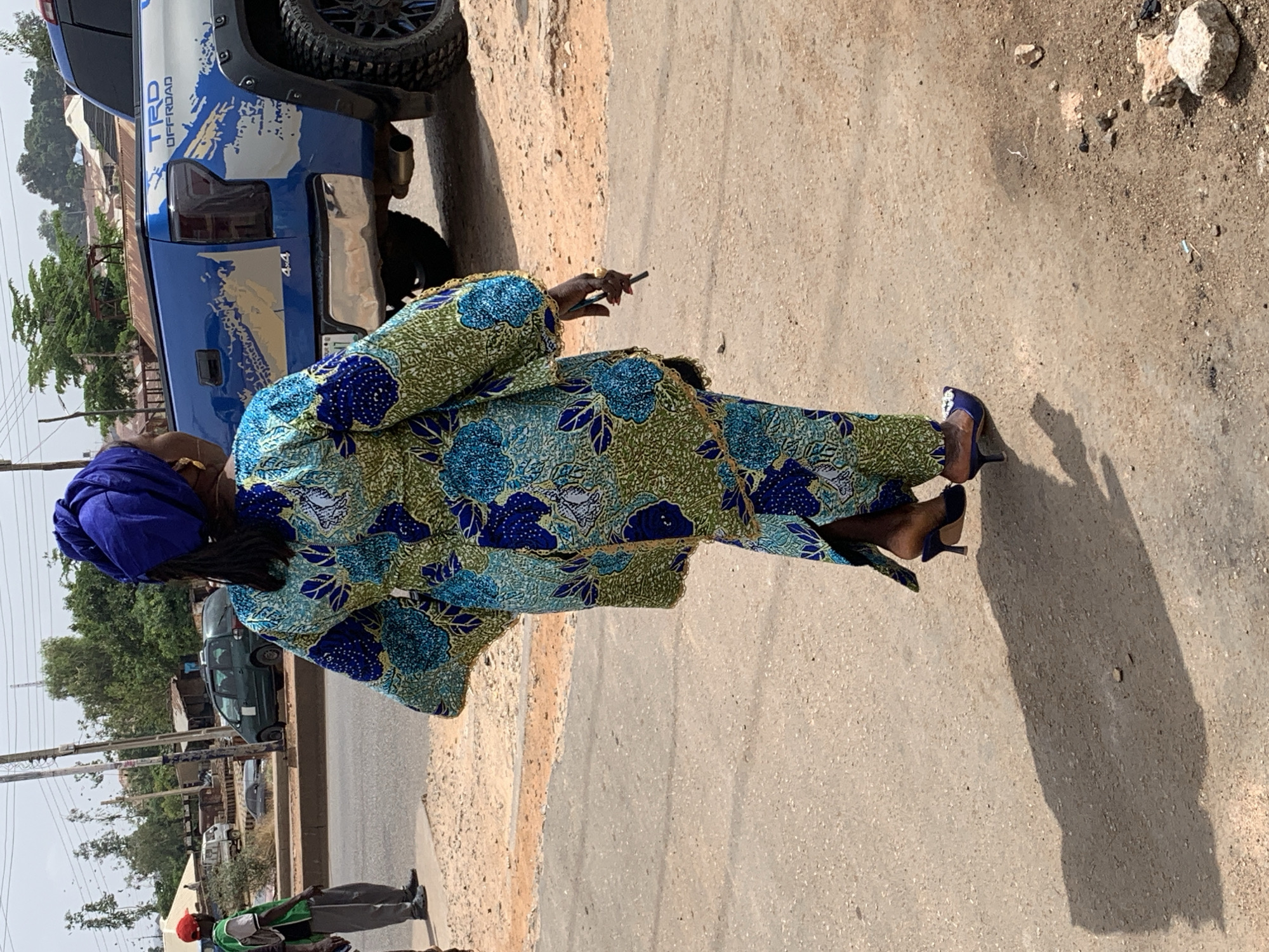 The image shows a woman in Bukuru, Nigeria, standing on a paved street. She is wearing a blue headwrap and a blue and green patterned outfit with blue high-heeled shoes. She is holding a thin, dark object in her hand. A blue pickup truck with "TRD Offroad" lettering is parked nearby. Another person is partially visible on the left side of the frame, wearing a red cap and a green shirt. The background includes buildings, trees, and utility poles. The street surface appears to be concrete or asphalt.