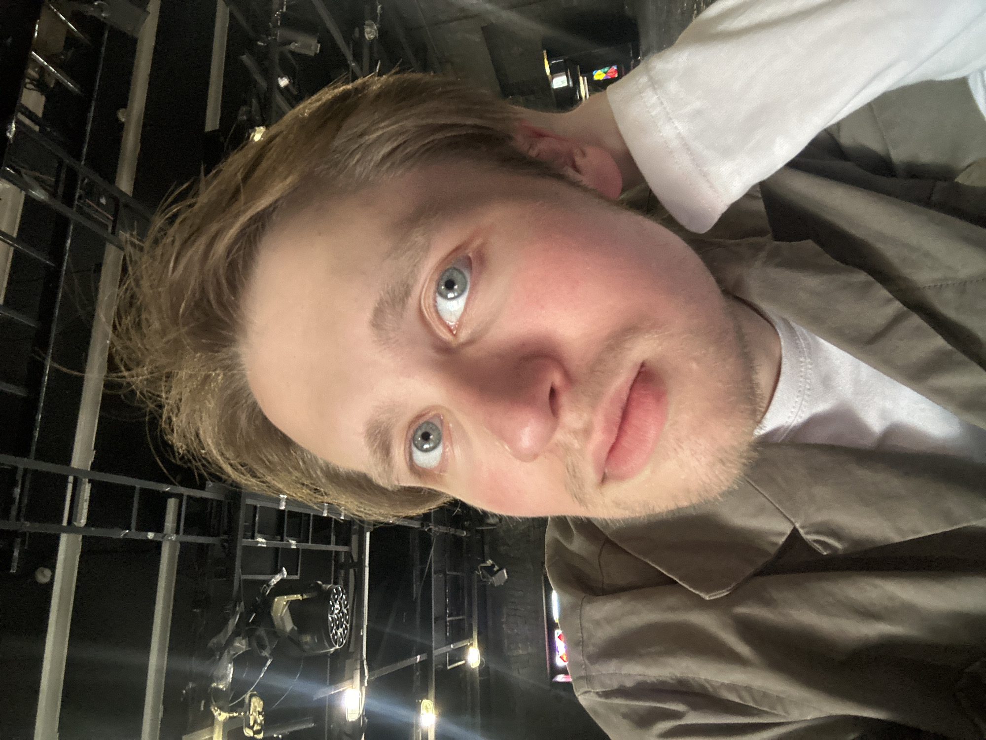 A close-up, portrait-style photograph shows a fair-skinned person with light blonde hair and blue eyes looking directly at the camera. The person is wearing a white t-shirt and a light brown or khaki jacket. The background features a dark, industrial-looking structure with metal bars and lighting fixtures. Some lights are shining brightly in the background. The photo appears to be taken in an indoor setting.