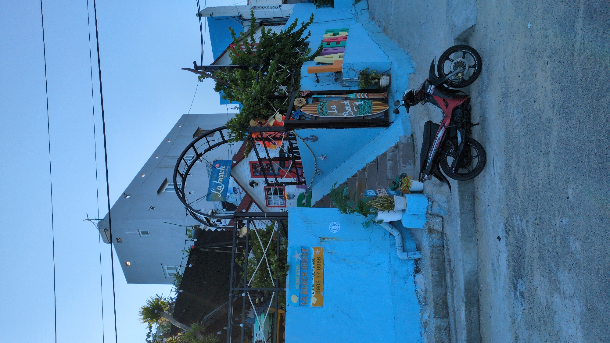 The image depicts a street scene in East Quy Nhon Ward, Vietnam, centered on a multi-tiered establishment. In the foreground, a dark red and black Yamaha scooter is parked on a grey concrete or asphalt surface, facing left. To the left, a vibrant blue concrete wall with a visible drainpipe runs along the base of a structure. A yellow rectangular sign, affixed to this blue wall, displays "HOMESTAY LA BEACH HOUSE NHON LY QUY NHON" and the phone number "0905 37 0086".
A set of concrete steps, painted blue on the sides, ascends from the left towards a landing. Potted plants in white containers are positioned at the base of the stairs. Above the stairs, a small, white, house-like structure with a red door and red-trimmed windows is visible, partially obscured by an arched black metal structure adorned with hanging spherical lights. A blue sign bearing "La beach Homestay - Cafe" hangs from this arch. Green foliage and various beach-themed decorations, including anchors, starfish, and oars, are present around this upper level. To the right, large, multi-colored block letters spelling "LA BEACH" are affixed to a blue wall, adjacent to a tall, vertical wooden sign resembling a surfboard, which also reads "La Beach" and the numeral "1".
In the background, behind the colorful establishment, stands a multi-story, light grey building with numerous small, rectangular windows. A rooftop structure, possibly an antenna or utility fixture, is visible at its apex. Overhead, several black utility lines traverse the clear, light blue sky. No individuals are present within the frame.