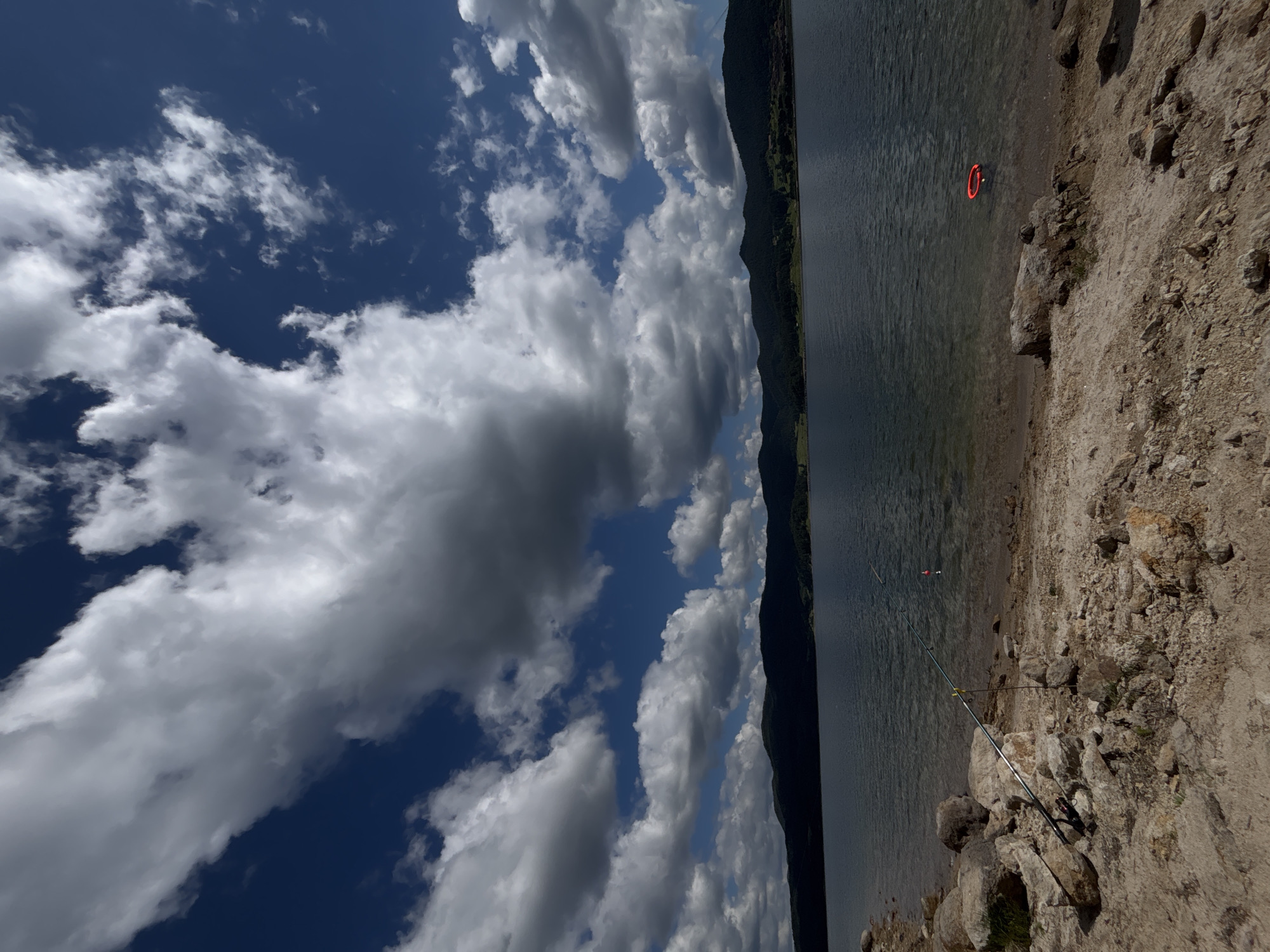 A wide-angle, low-angle shot depicts a scenic view of a serene lake on a partially cloudy day. The foreground is dominated by a rocky, sandy shore. Two fishing rods are set up on the shore, one in the left foreground and another further back, with a red float visible on the water. In the lake, a bright red, circular object, possibly a flotation device, is floating near the shore. The middle ground features the calm, dark blue water of the lake. The background showcases a chain of dark, silhouetted mountains covered in lush green trees, with patches of lighter green suggesting fields or settlements. The sky above is a vibrant blue, streaked with dramatic, large, white and gray clouds that cast shadows on the landscape, creating a dynamic contrast between light and dark. The overall atmosphere is peaceful and natural, with a sense of solitude. The scene is located in Unknown, Unknown.