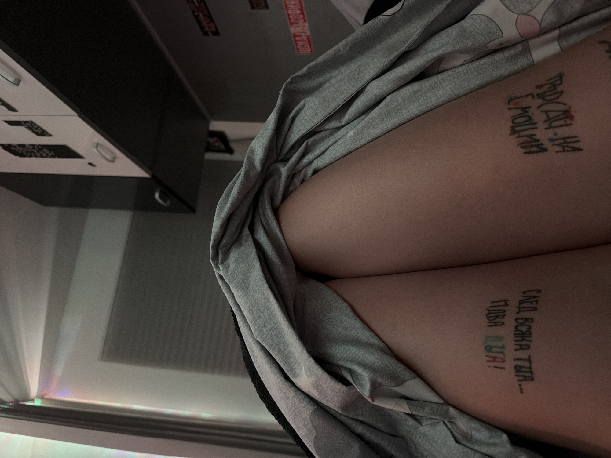 The image shows a close-up of a person's thighs, covered in part by a grey patterned cloth. Each thigh has a black tattoo in Cyrillic. The background includes a dark cabinet or shelf and a window with a light source. The location is not identifiable as being in Varna, Bulgaria.