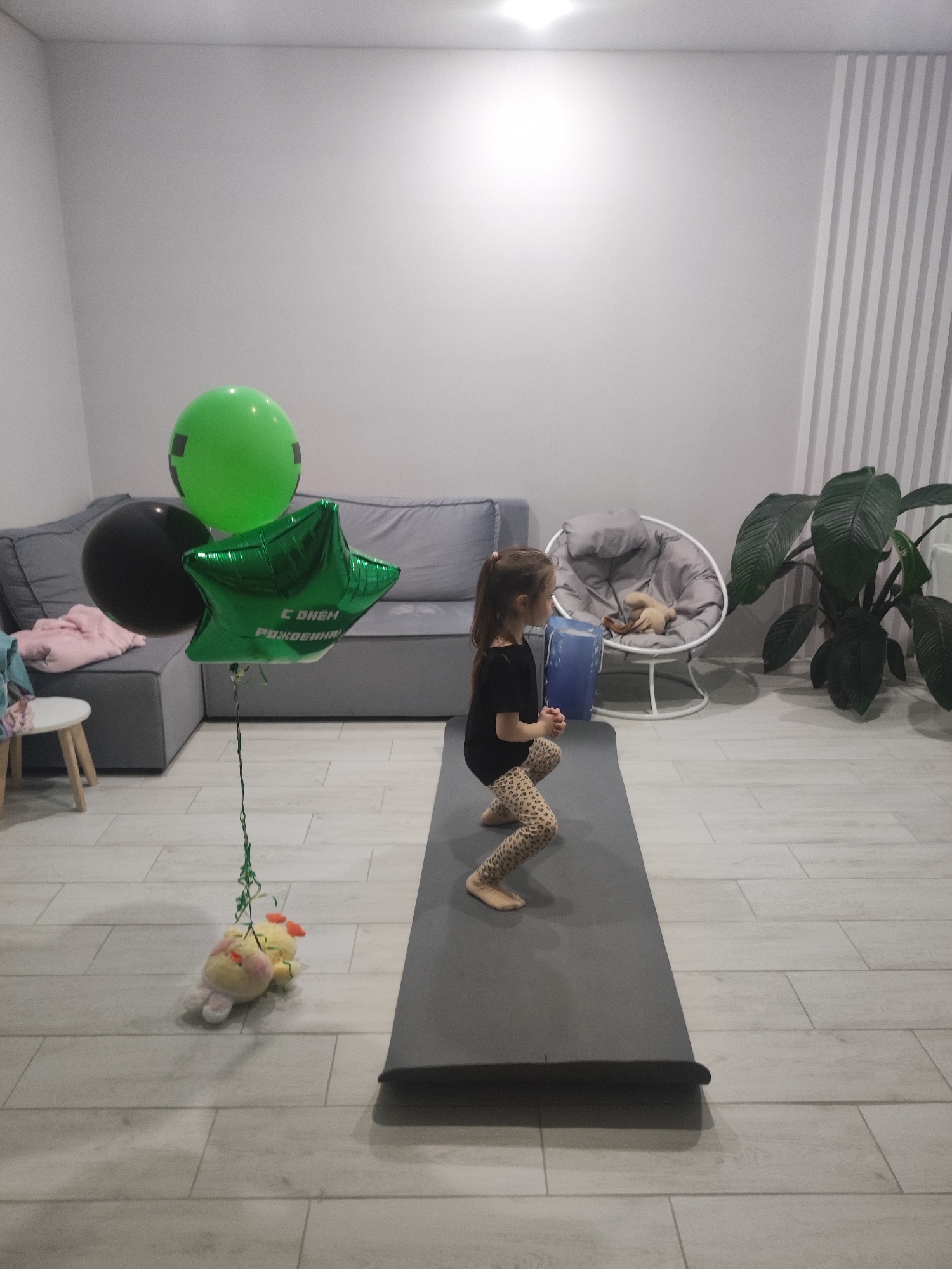 A young girl is exercising on a gray yoga mat in a room. She is wearing a black shirt and leopard-print leggings and is in a squat position holding a blue object. To her left are three balloons: a green balloon, a black balloon, and a green star-shaped balloon with text in Cyrillic that says "С ДНЕМ РОЖДЕНИЯ!" (Happy Birthday!). Attached to the balloon strings is a plush toy. Behind the girl, there is a gray sofa, a round chair with a light gray cushion and a plush toy on it, and a large potted plant. The room has light-colored tiled flooring and white walls, one of which has vertical grooves. The location is likely a home or apartment, but without further information, it is impossible to confirm it is in Votkinsk, Russia.